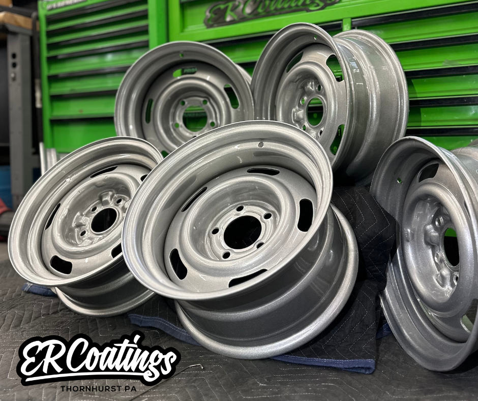 Several silver-painted steel vehicle wheels stacked on a padded work surface in front of a green workshop tool chest.