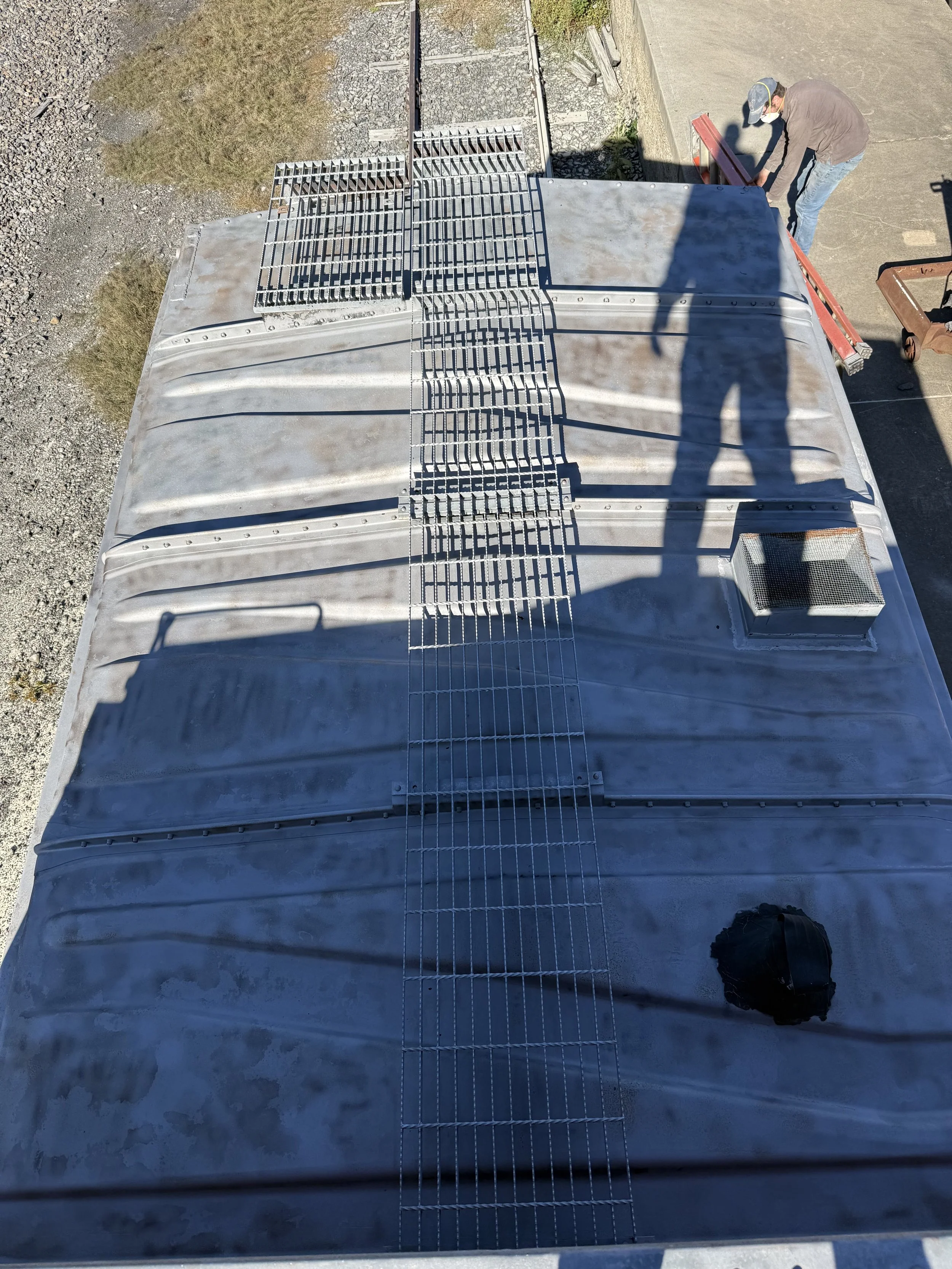 View from above of a metal roof with a vent, metal grates, and two workers installing or repairing on the roof. One worker is near the edge and the other is working on a scaffold.
