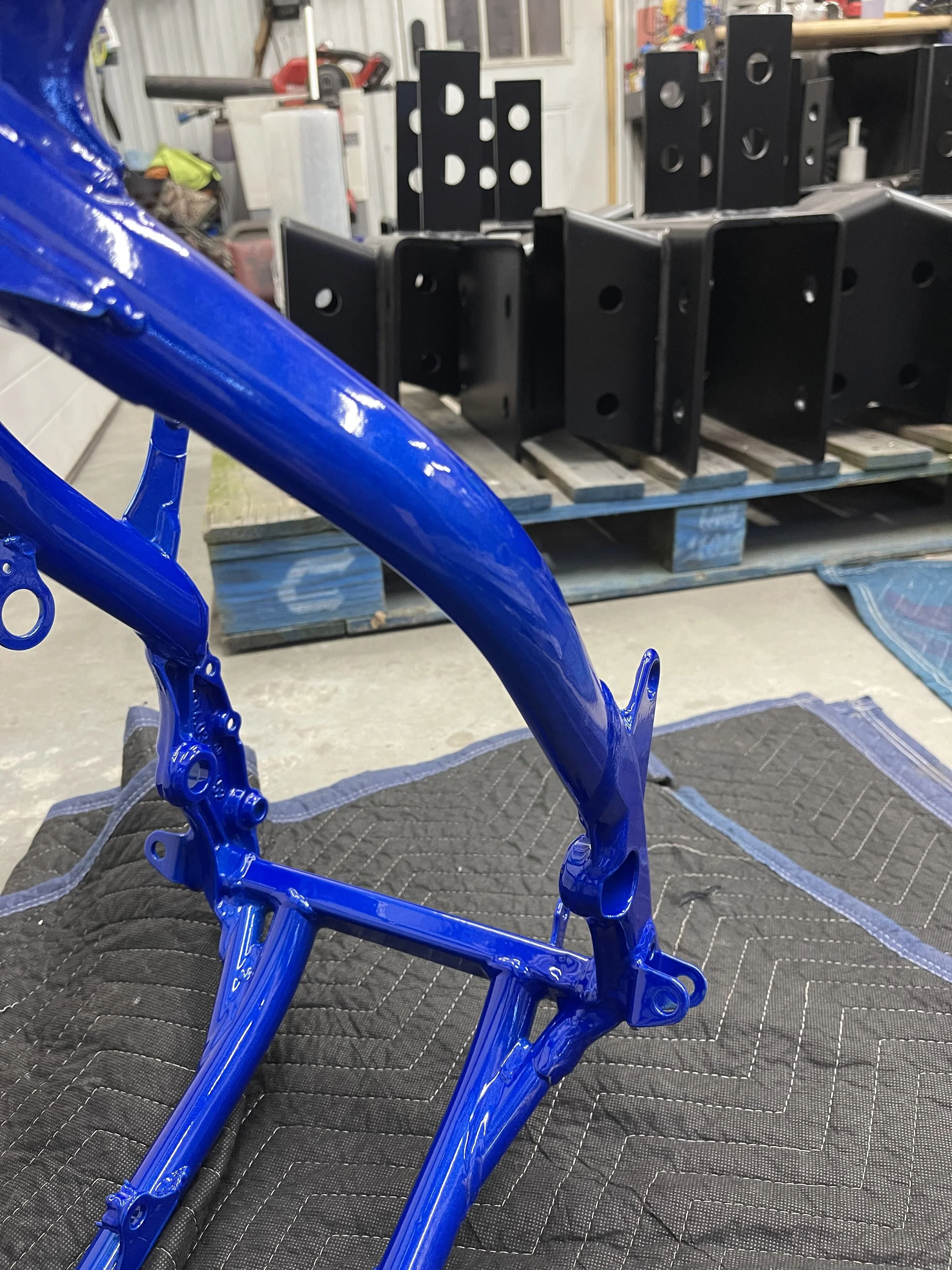 Blueprinted blue motorcycle frame on a padded work surface in a workshop, with black metal components and tools in the background.