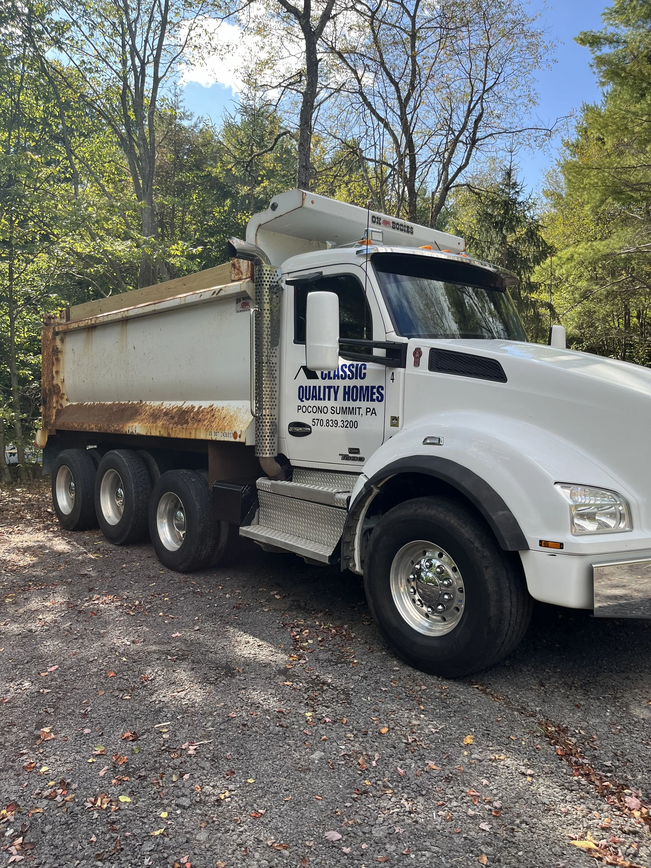 White dump truck with rusty bed parked on a wooded area, with a sign that reads 'Classic Quality Homes, Pocono Summit, PA, 570.839.3200'.
