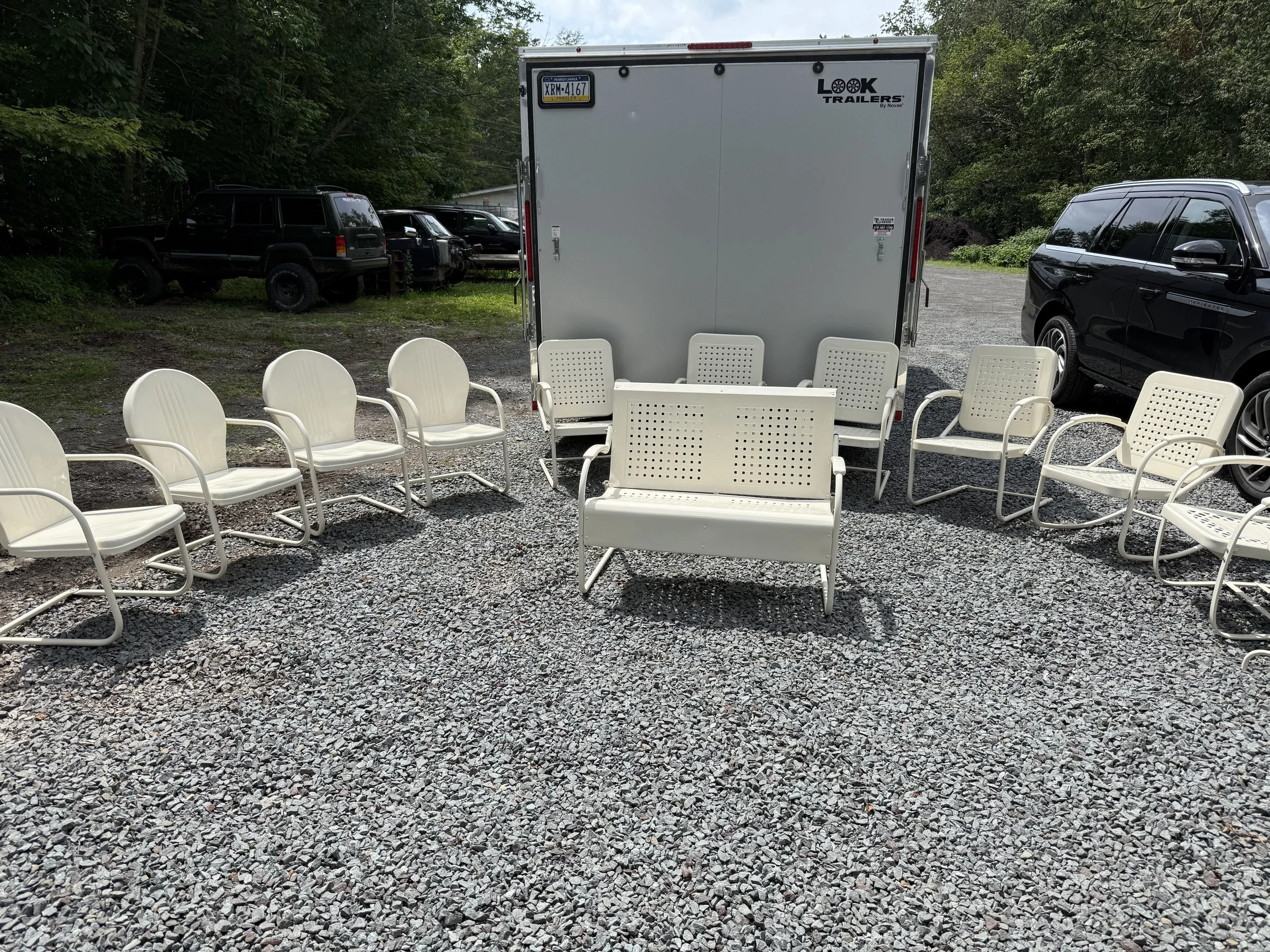 Outdoor scene with white chairs and a bench arranged in a semi-circle on gravel, with a white trailer in the background and parked cars on either side.