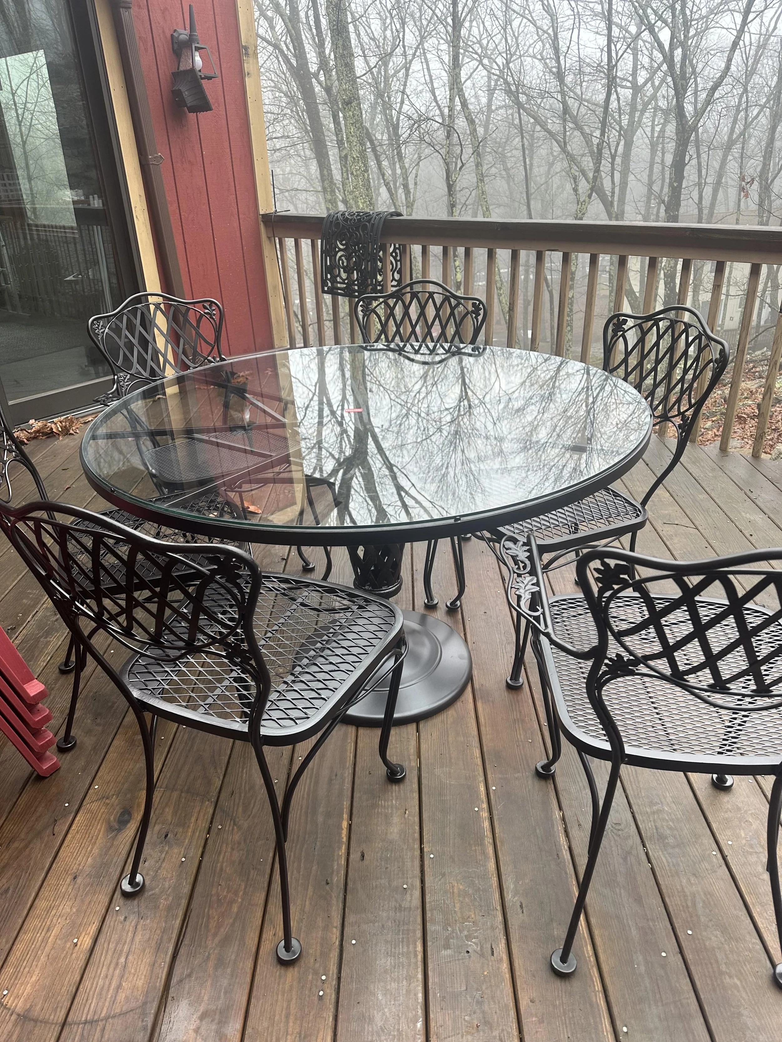 Outdoor wooden deck with a round glass table and six black metal chairs, surrounded by leafless trees and a foggy background.