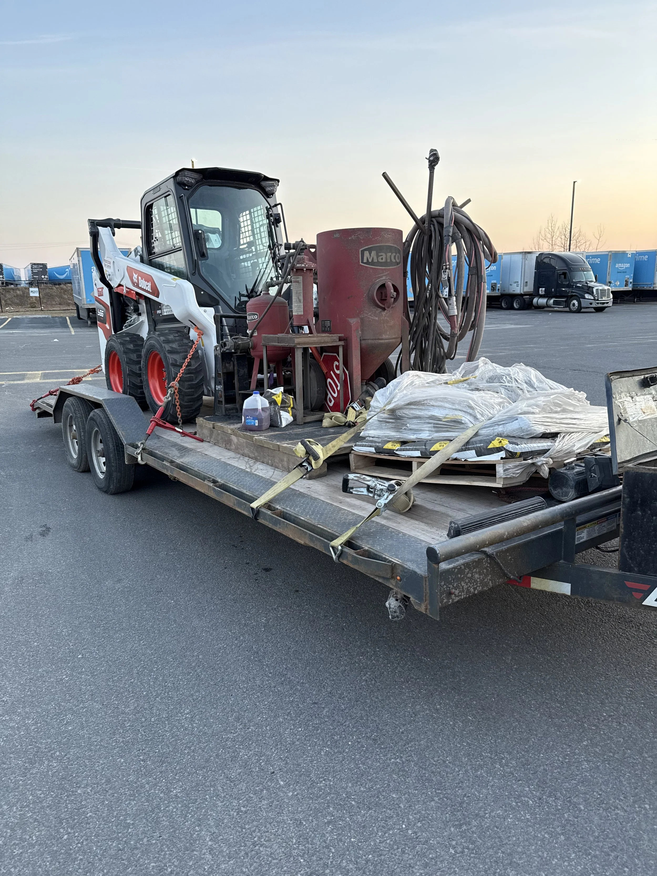 Flatbed trailer with construction equipment and materials, including a Bobcat skid-steer loader, hoses, a large red Marco container, and bags of supplies, parked in an outdoor lot during dusk.