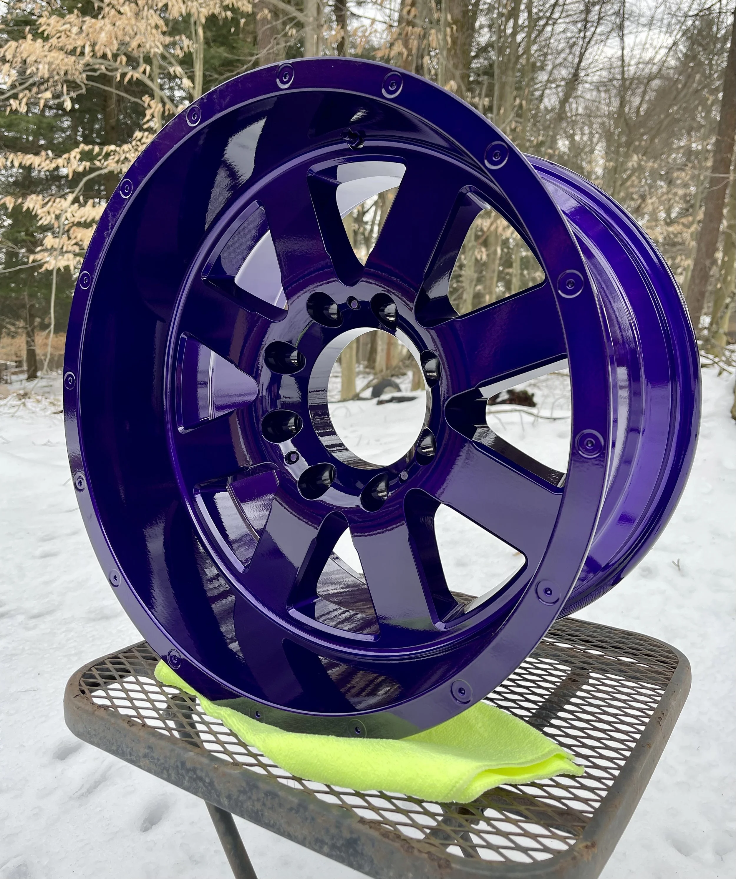 Purple alloy wheel rim with ten spokes resting on a yellow cloth on a metal table outdoors in a snow-covered area with trees in the background.
