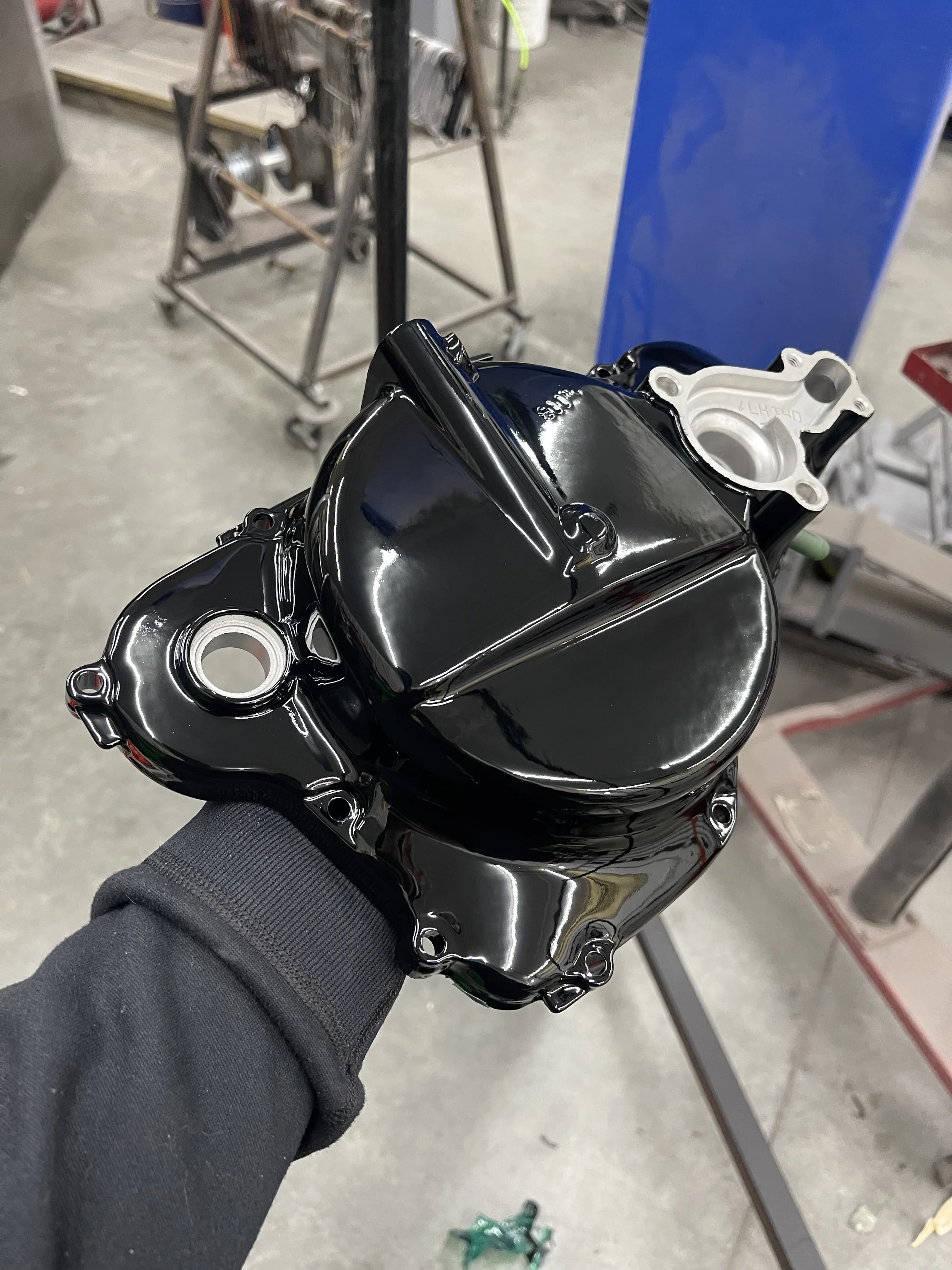 A person holding a shiny black engine cover in a workshop, with tools and equipment in the background.