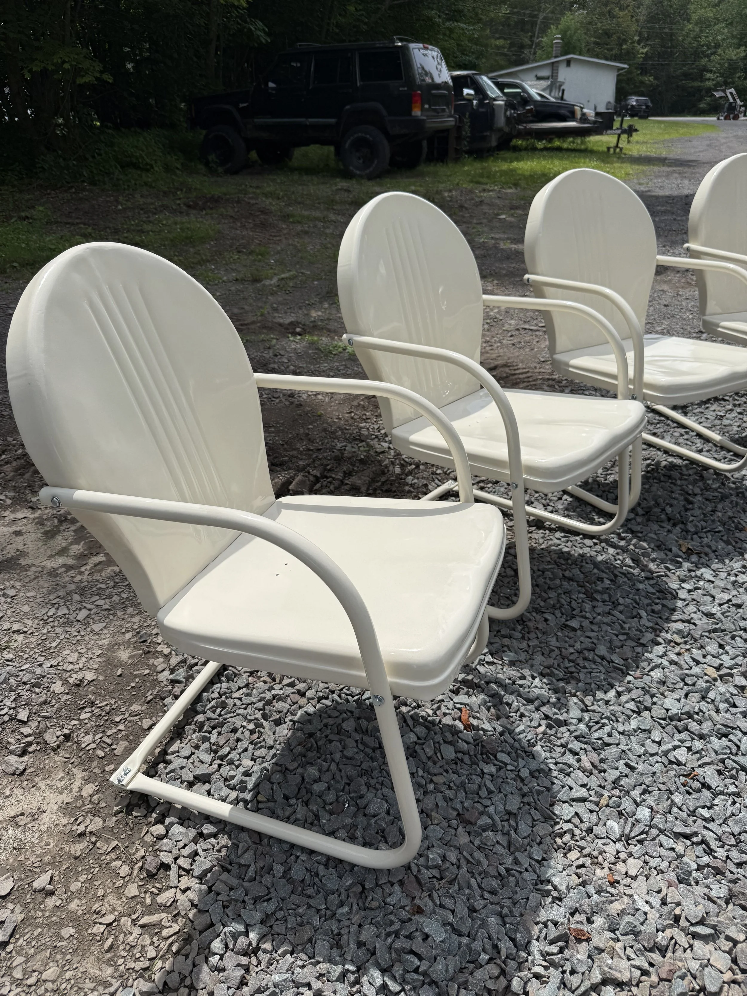 Three white outdoor chairs with armrests arranged on gravel ground in a row.