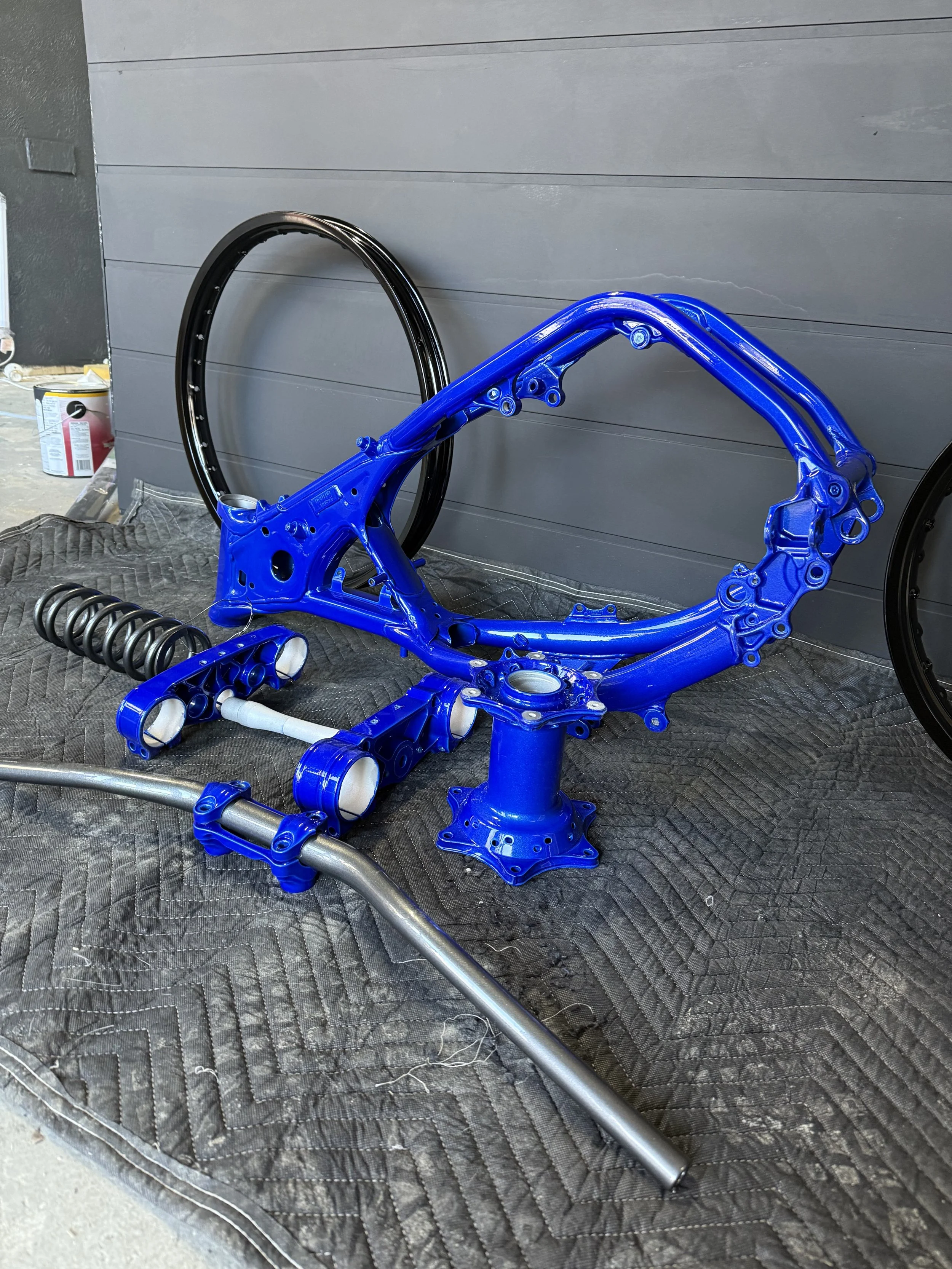 Disassembled blue motorcycle frame with a wheel, spring, and handlebar parts on a padded mat.