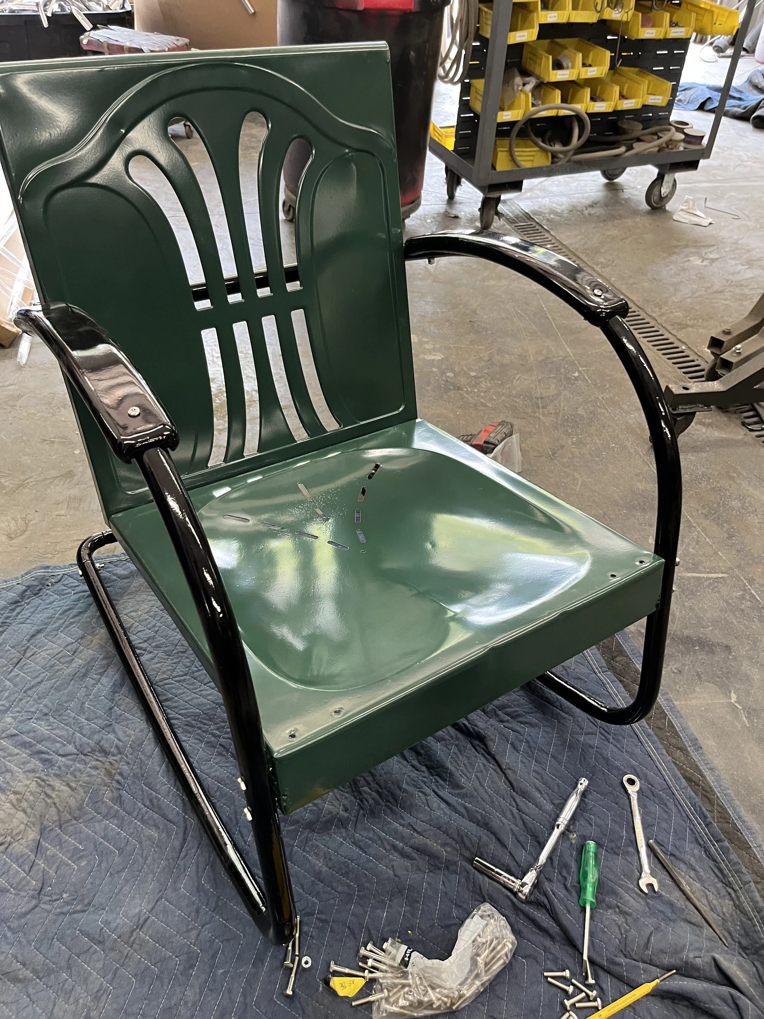 Disassembled green and black metal outdoor chair on a blanket, with tools and hardware around it in a workshop.