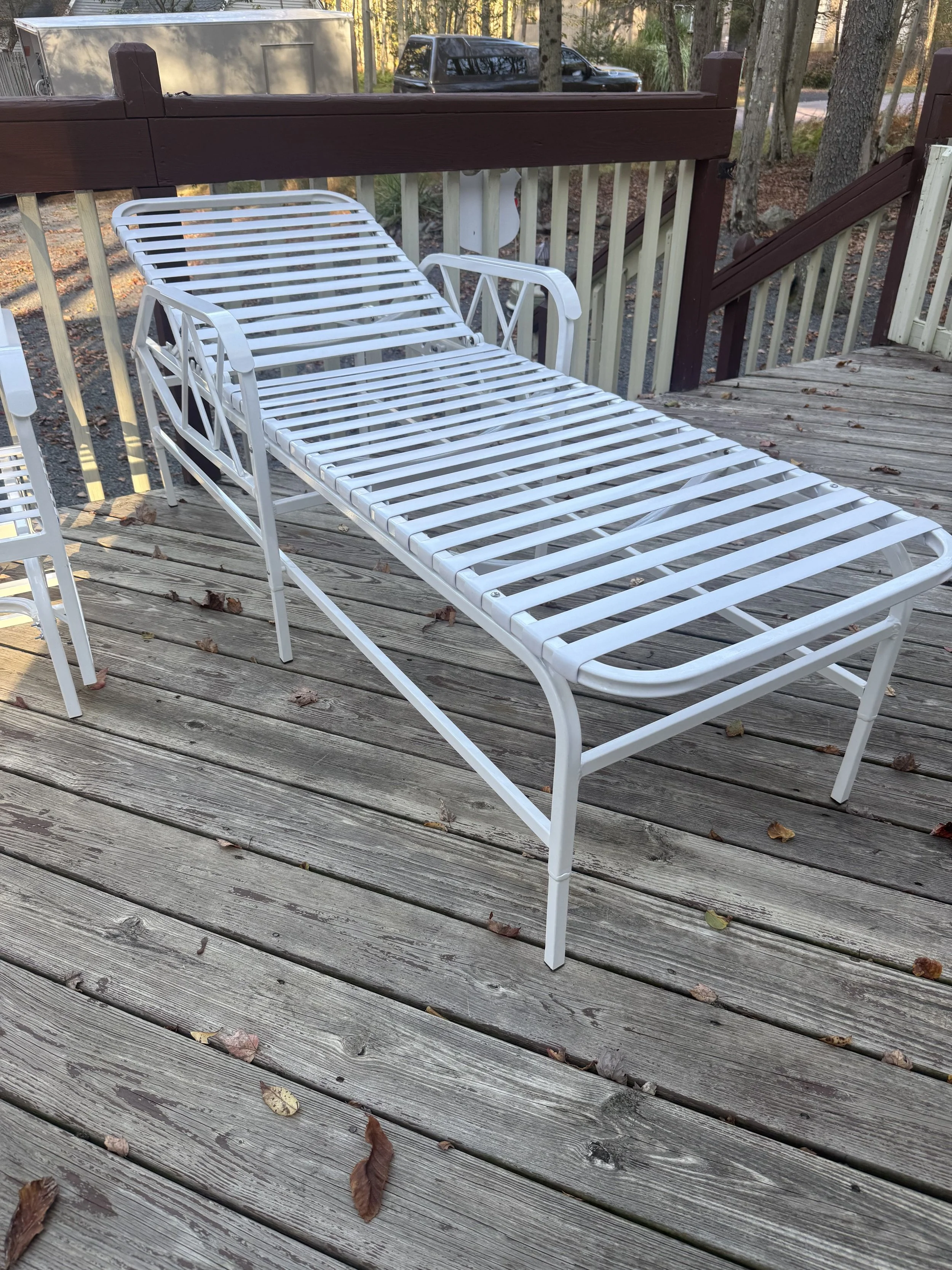 A white metal outdoor lounge chair on a wooden deck, with fallen leaves scattered on the deck, and trees and a parked vehicle in the background.