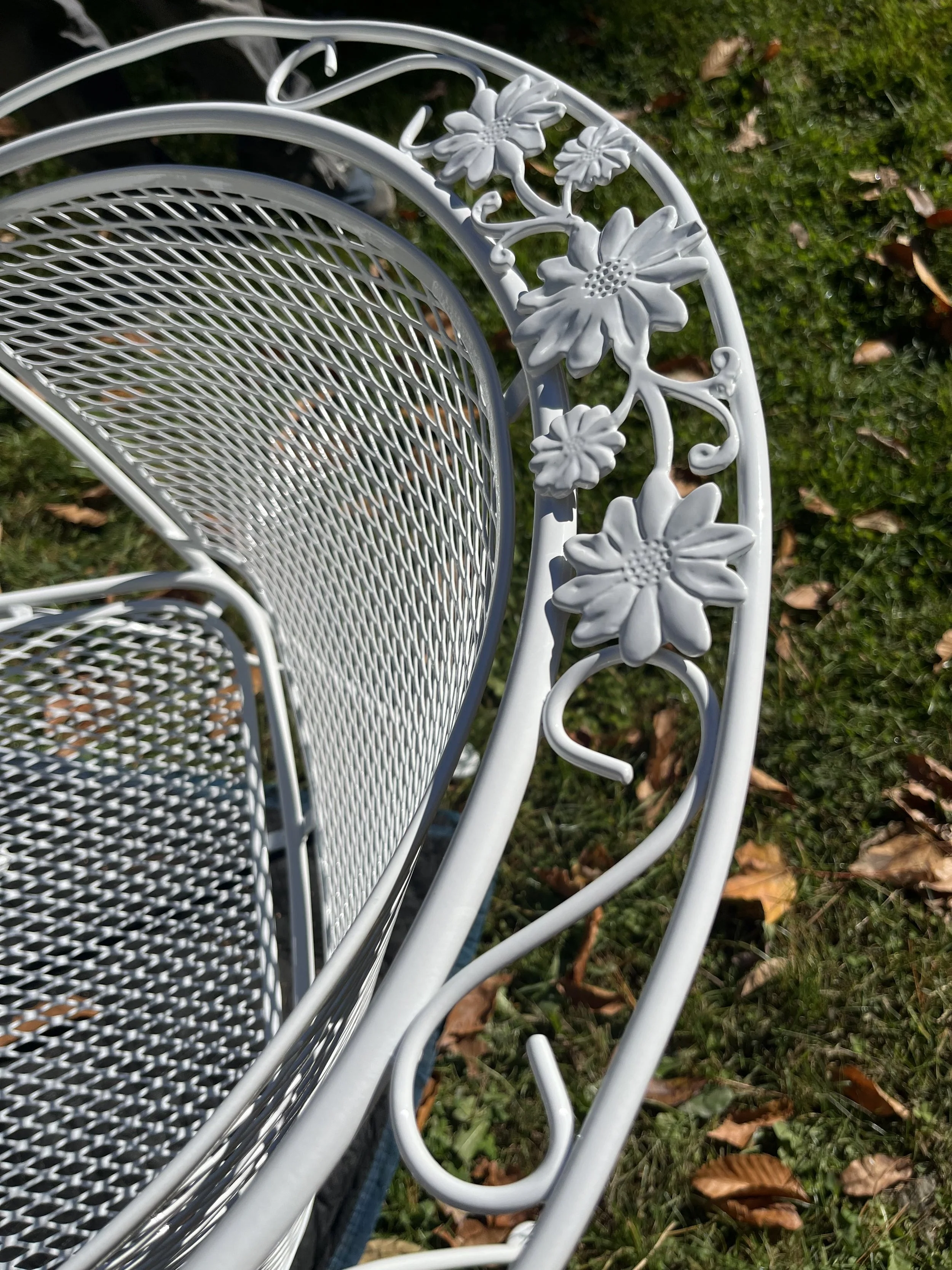 Close-up of a white metal outdoor garden bench with floral design details on the armrest, placed on grass with fallen leaves.