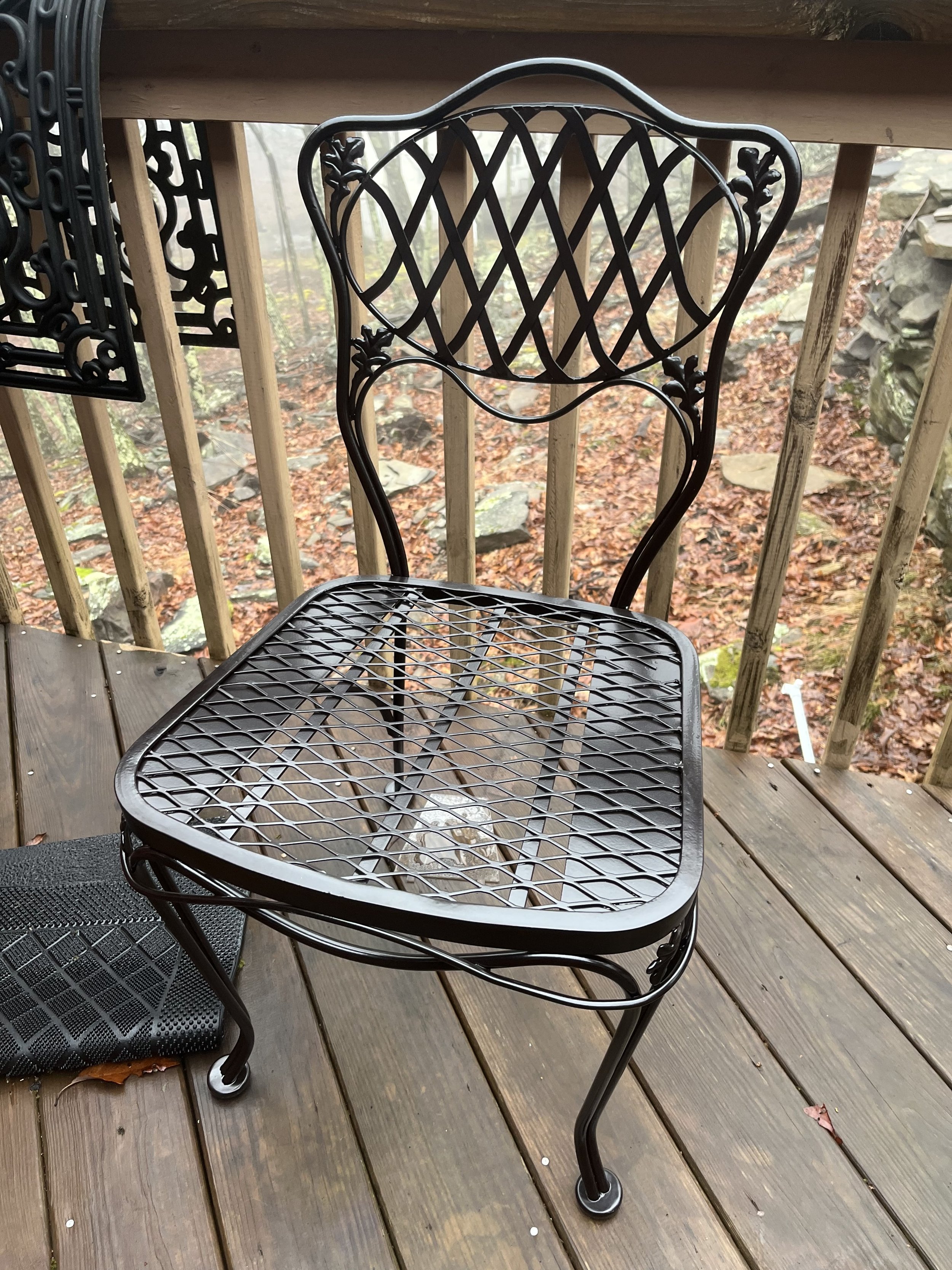 A black metal outdoor chair with a decorative backrest and a mesh seat, positioned on a wooden deck with a railing overlooking a leaf-covered yard.