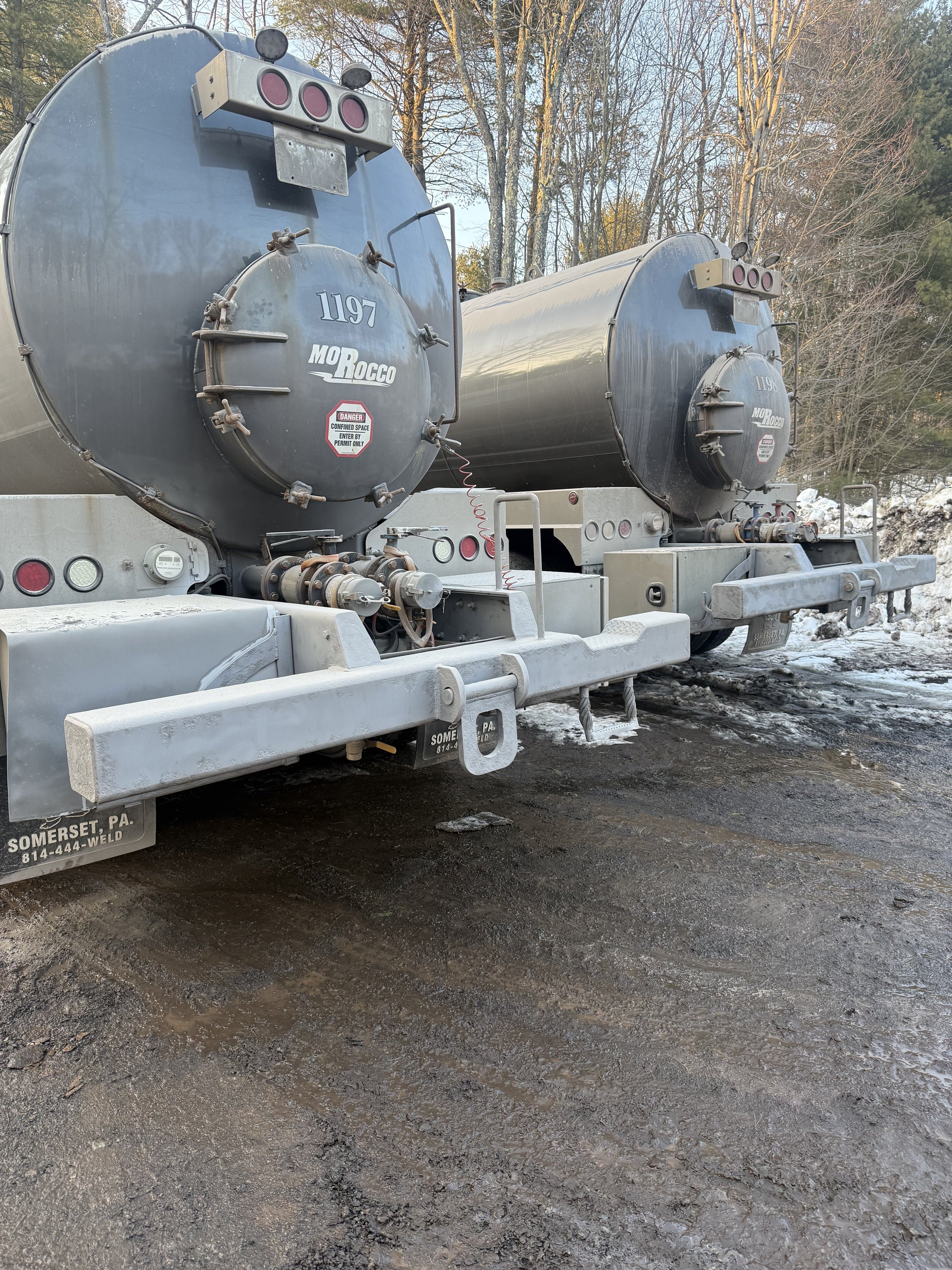 Two large gray tanker trucks parked on dirt ground with trees in the background.