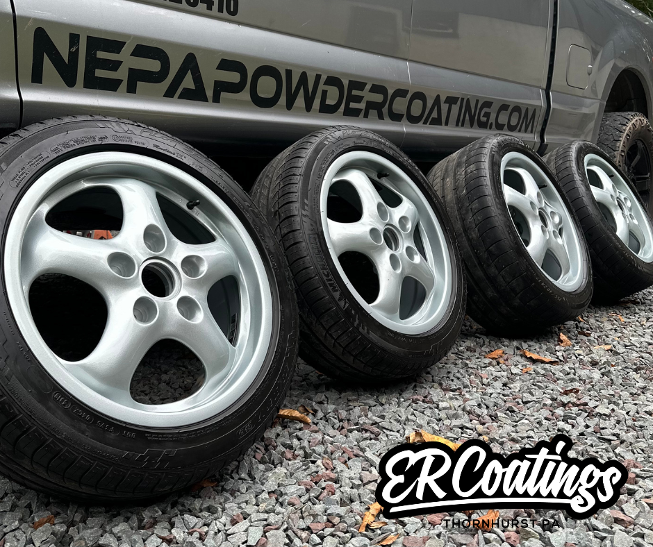 Four car tires with silver rims lined up on gravel in front of a vehicle with a banner reading 'NEPA POWDERCOATING.COM' and a logo 'ERC Coatings THORNHURST PA'.