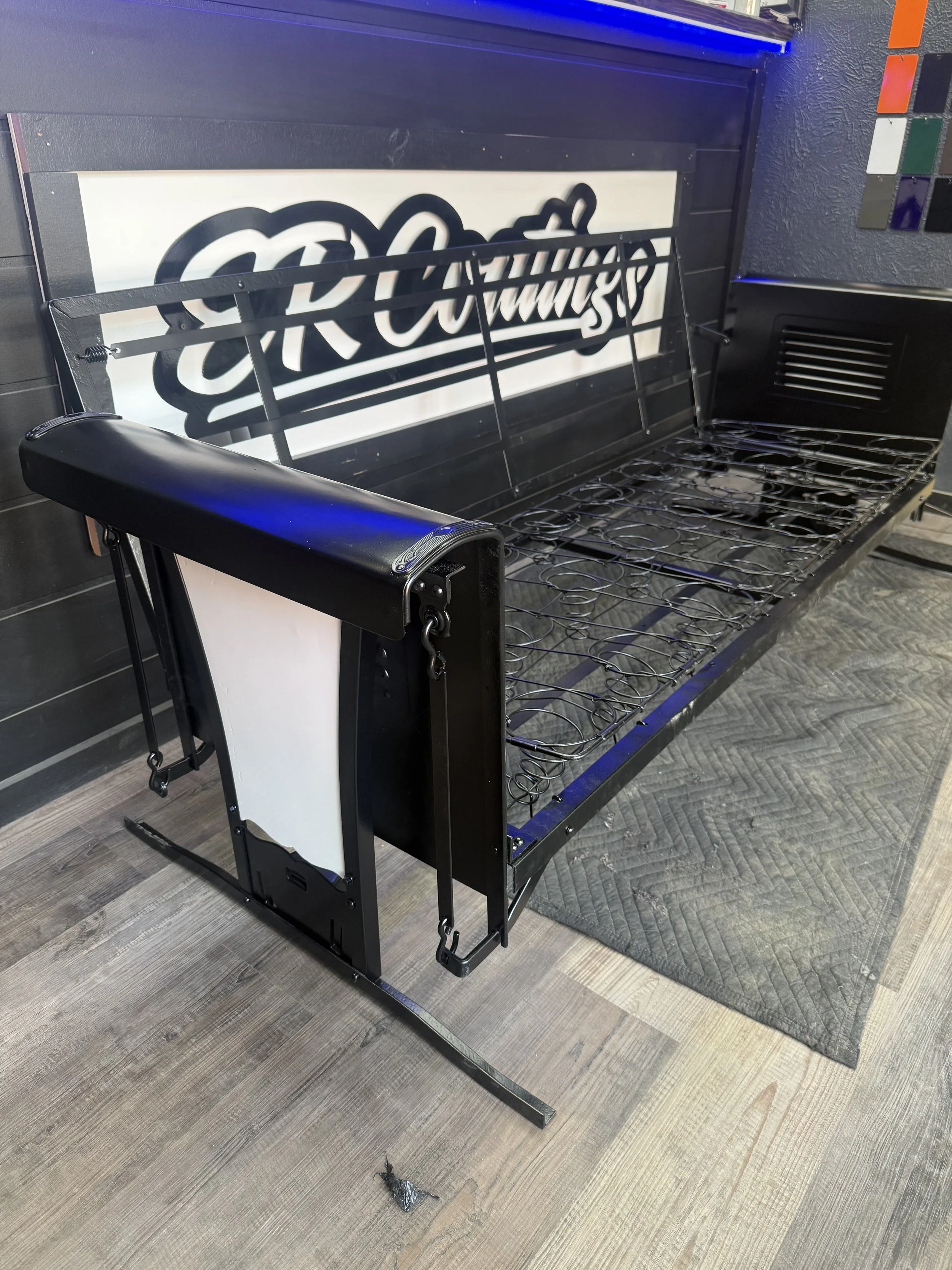 Empty black metal outdoor bench with a metal frame, placed on a wooden floor and a gray mat, in front of a black wall with a large white sign, in a commercial or retail space.