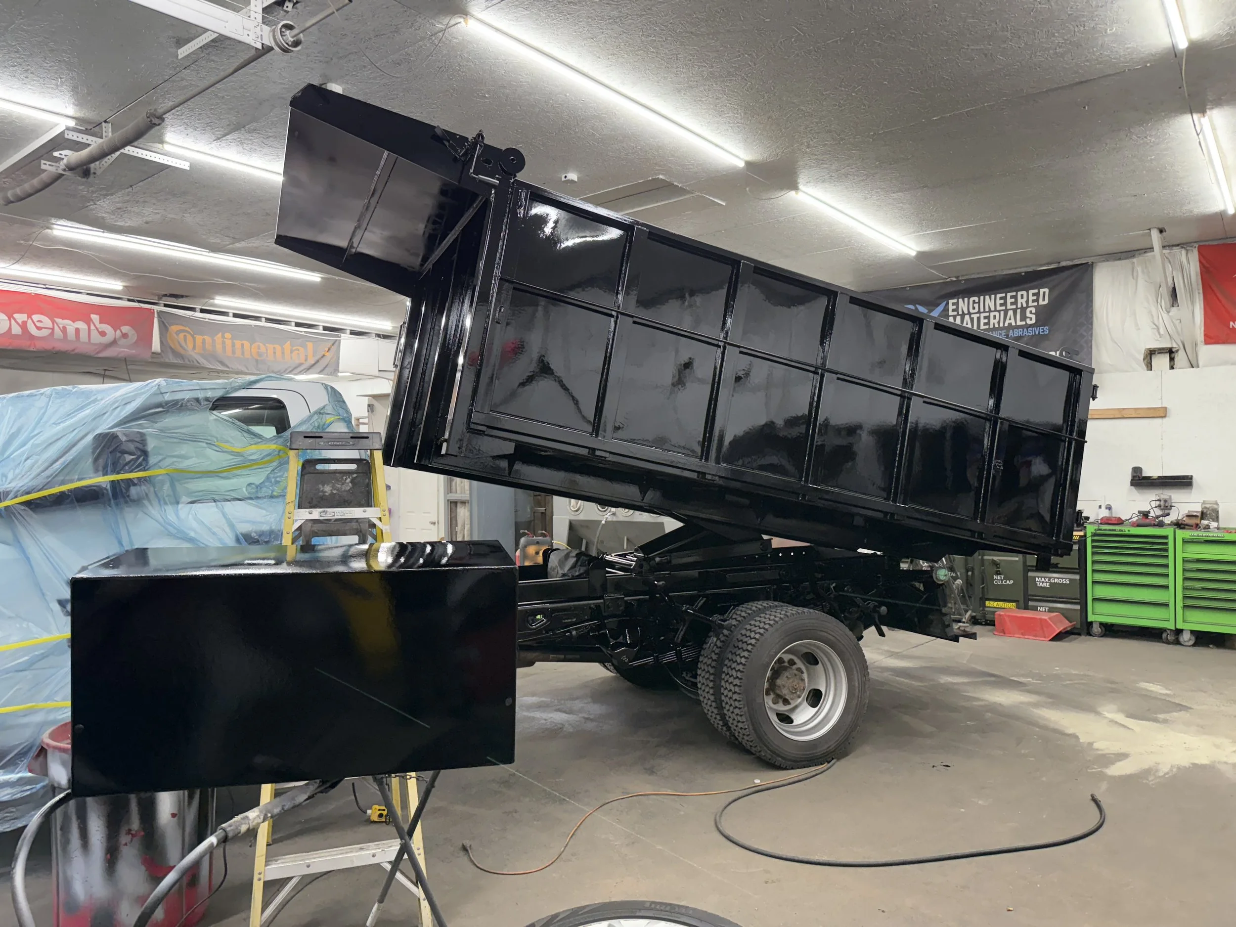 Black dump truck with its bed raised inside a workshop, with green tool chest, workbench, and supplies visible in the background.