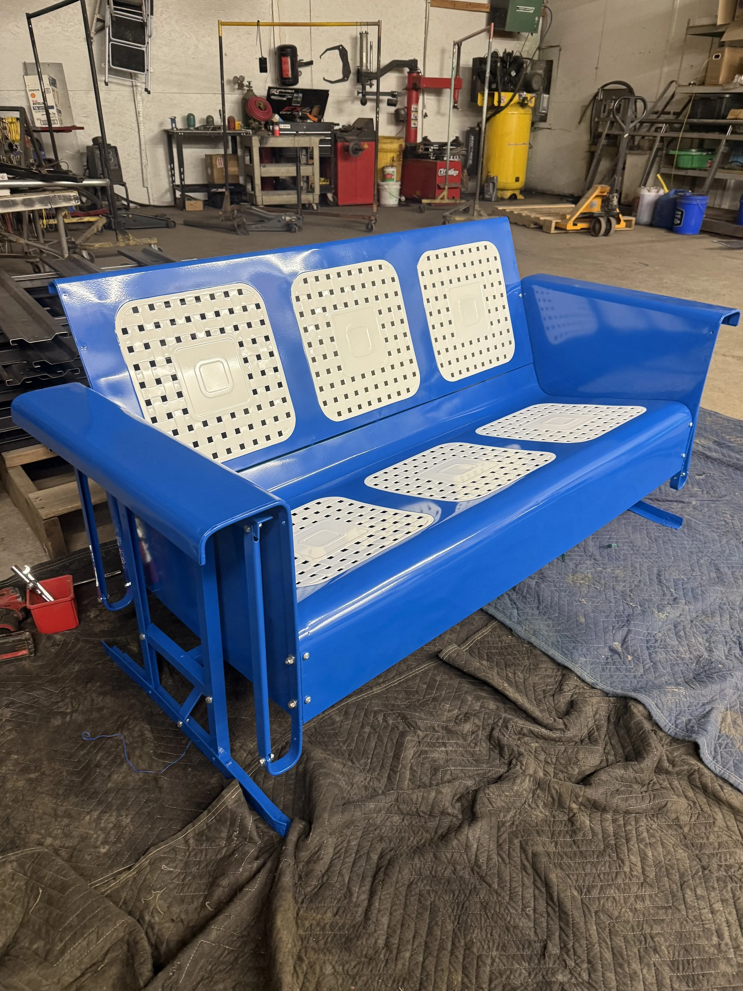 Blue metal bench with white perforated plastic seat and backrest in a workshop with various tools and equipment.