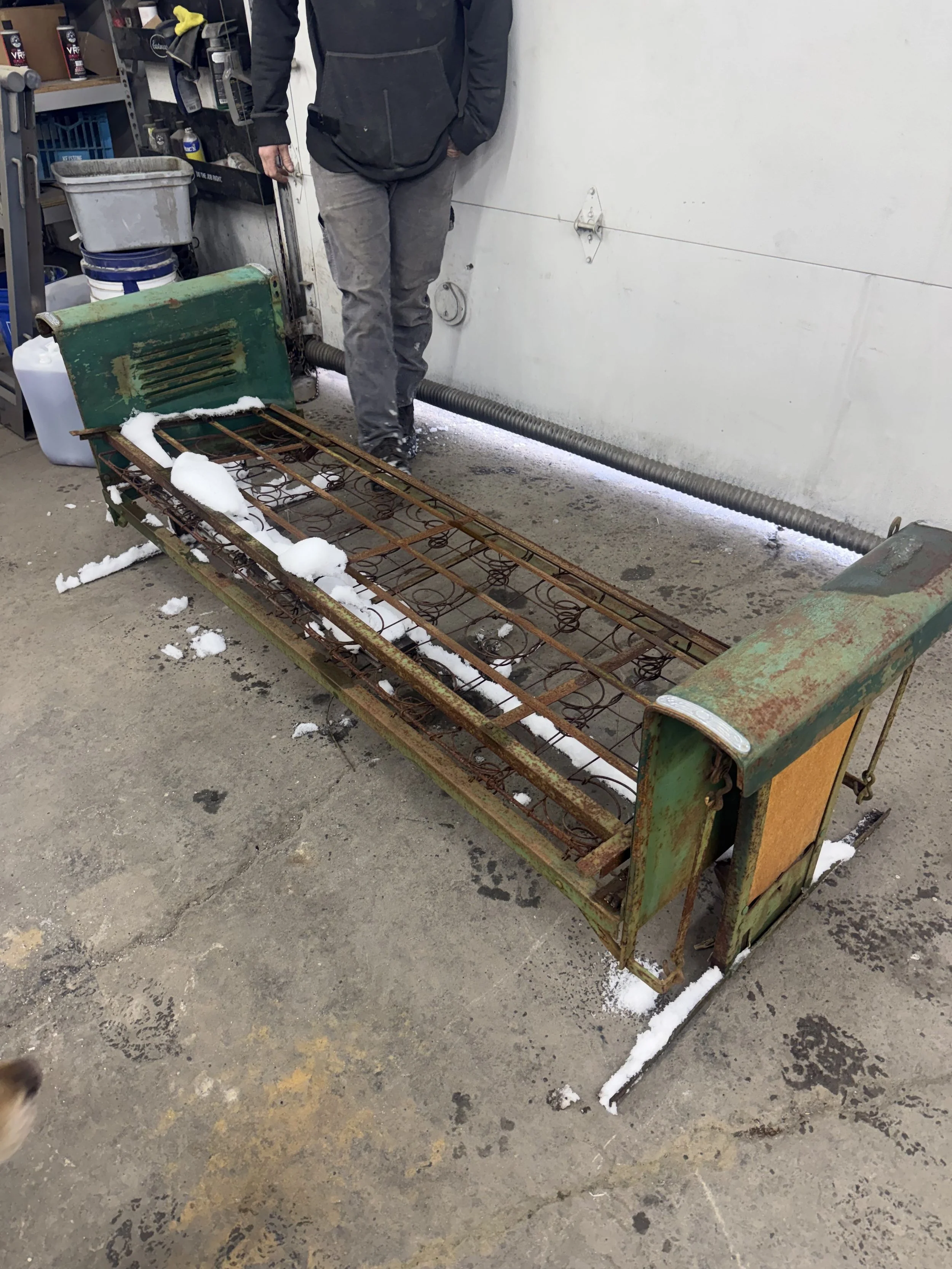 A rusty metal bed frame with snow on it, placed on a concrete floor in a workshop or garage. A person in black and gray clothing stands behind it. Shelves with various items are visible in the background.