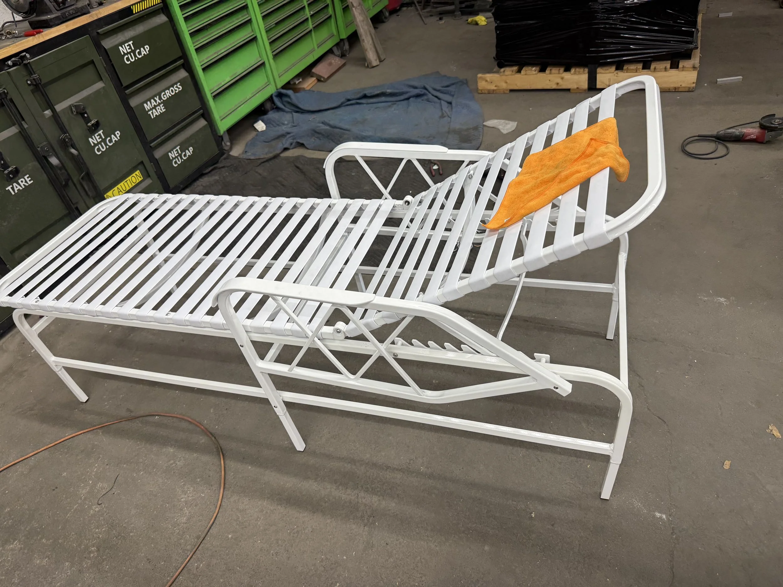 A white metal hospital gurney with an orange towel draped over the headrest, situated in a workshop or storage area with green tool chests, a blue cloth, and a power tool on the floor.