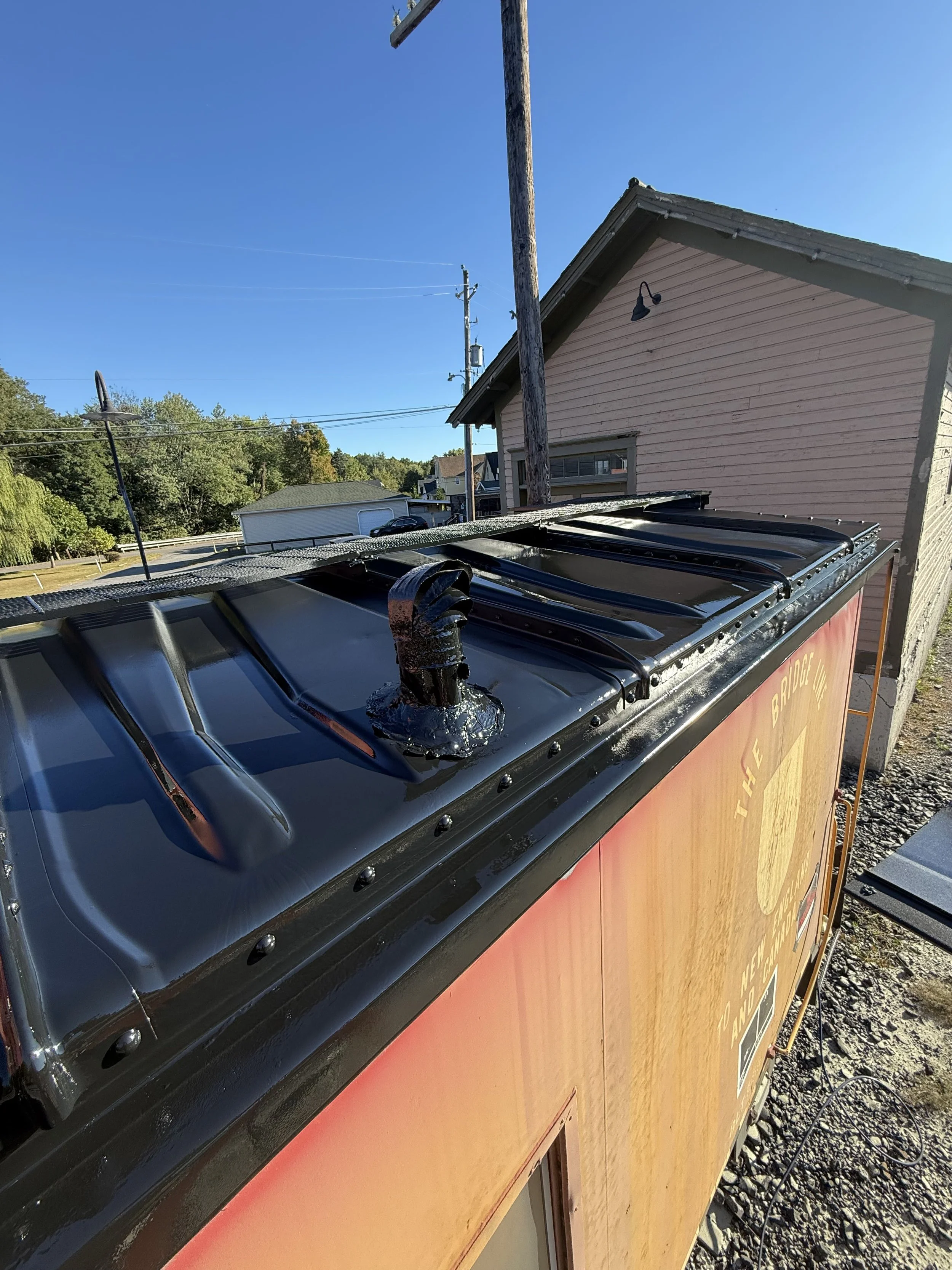 Close-up of a shiny black rooftop with vents, atop a red and yellow wooden trailer or small building, outside with blue sky and trees in the background.