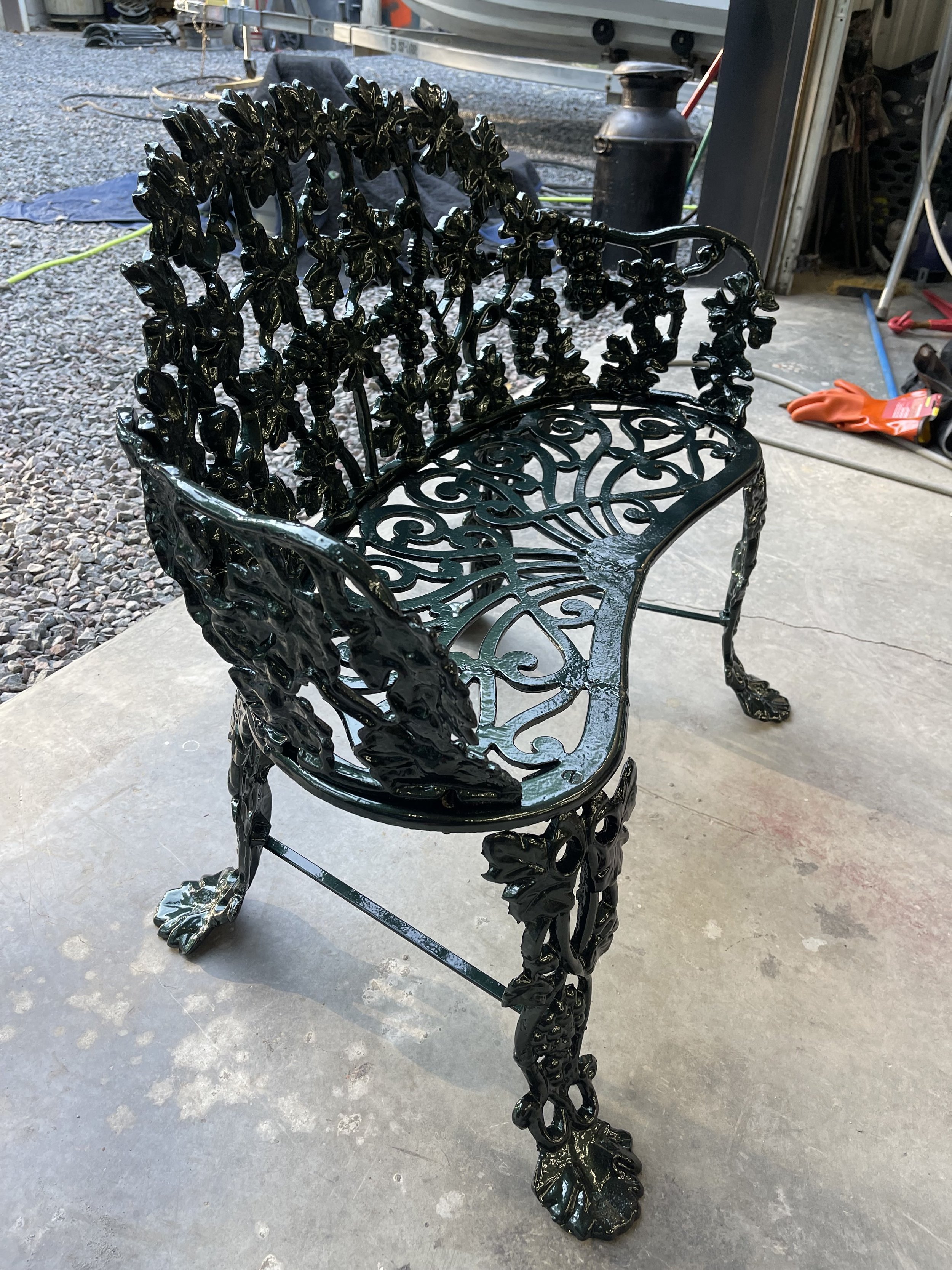 A black metal bench with ornate leaf and vine patterns, featuring lion paw feet, placed on a concrete patio with a workshop and gravel in the background.