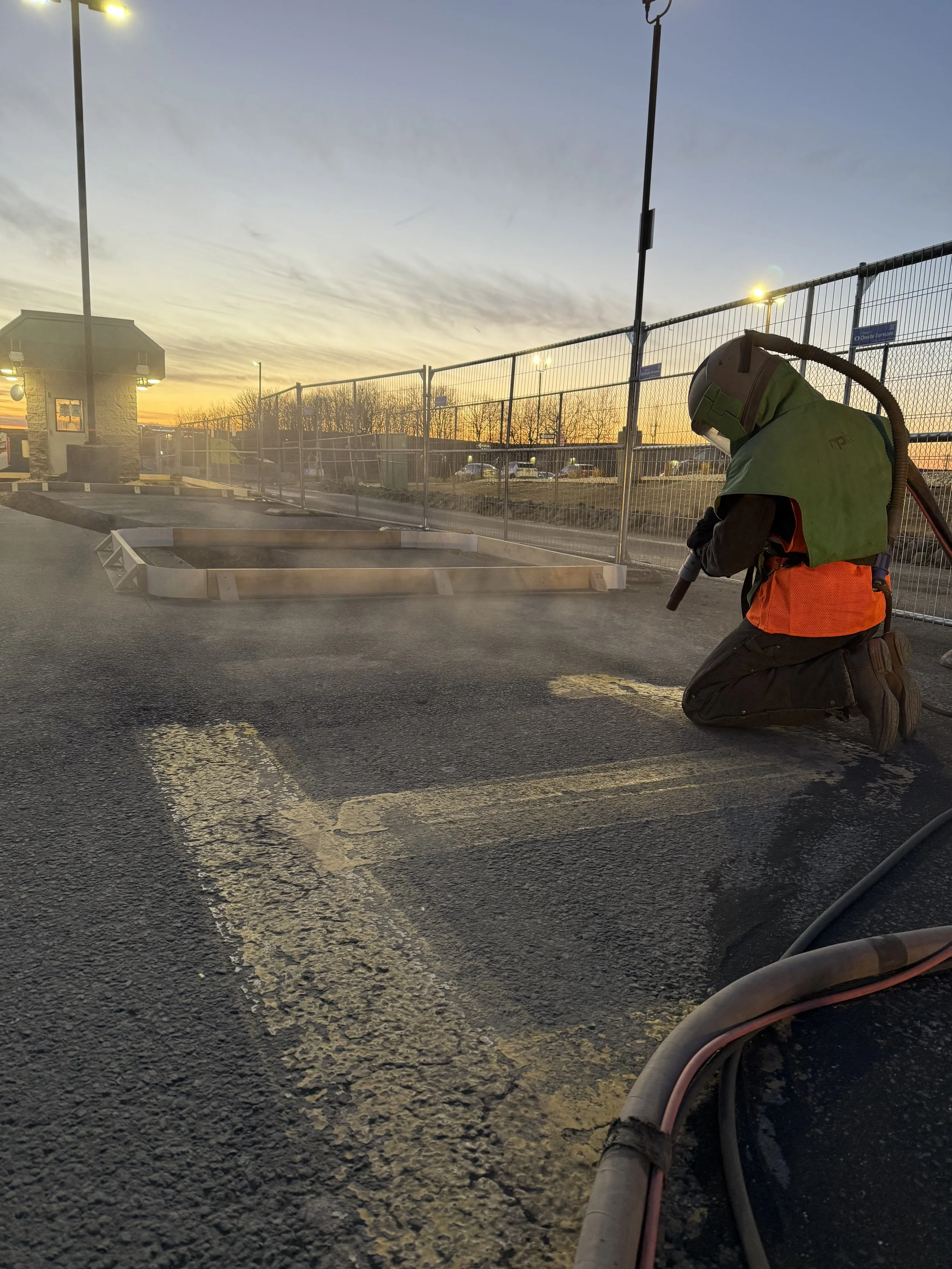 Worker kneeling on asphalt in a parking lot, welding or grinding a line on the ground during sunset with an orange safety vest and helmet, behind a chain-link fence.