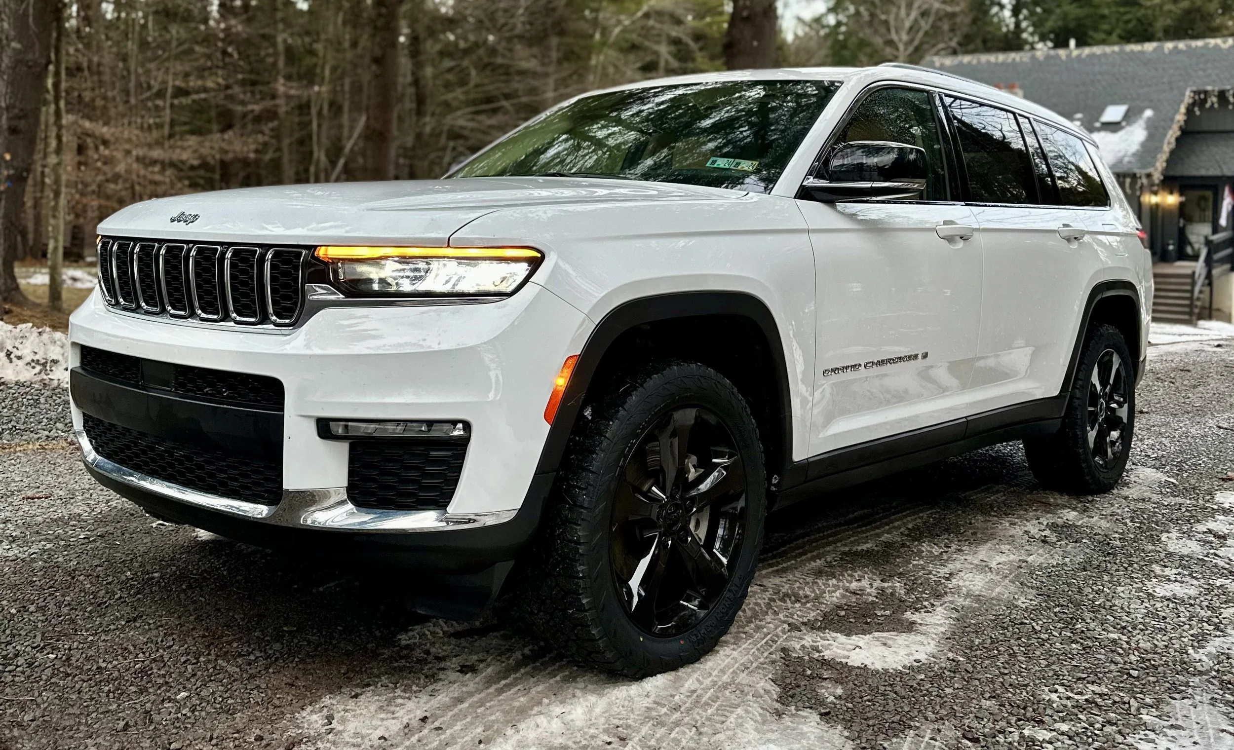 A white Jeep Grand Cherokee SUV parked on a snow-dusted gravel driveway in front of a wooded area with a house in the background.