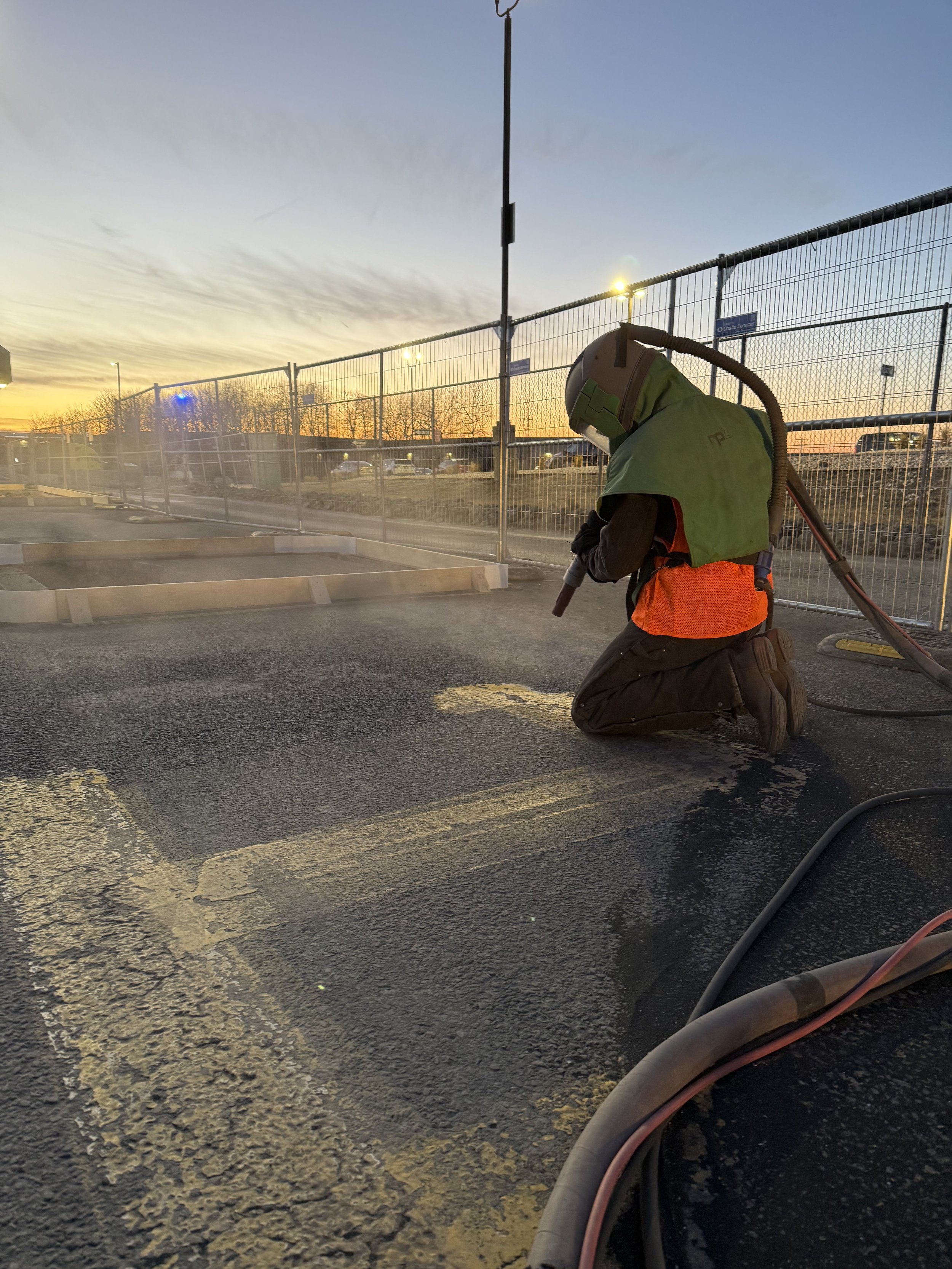 A construction worker kneels on the ground in a parking lot during sunset, wearing safety gear including a helmet, high-visibility vest, and kneepads, using a tool to work on the asphalt surface.