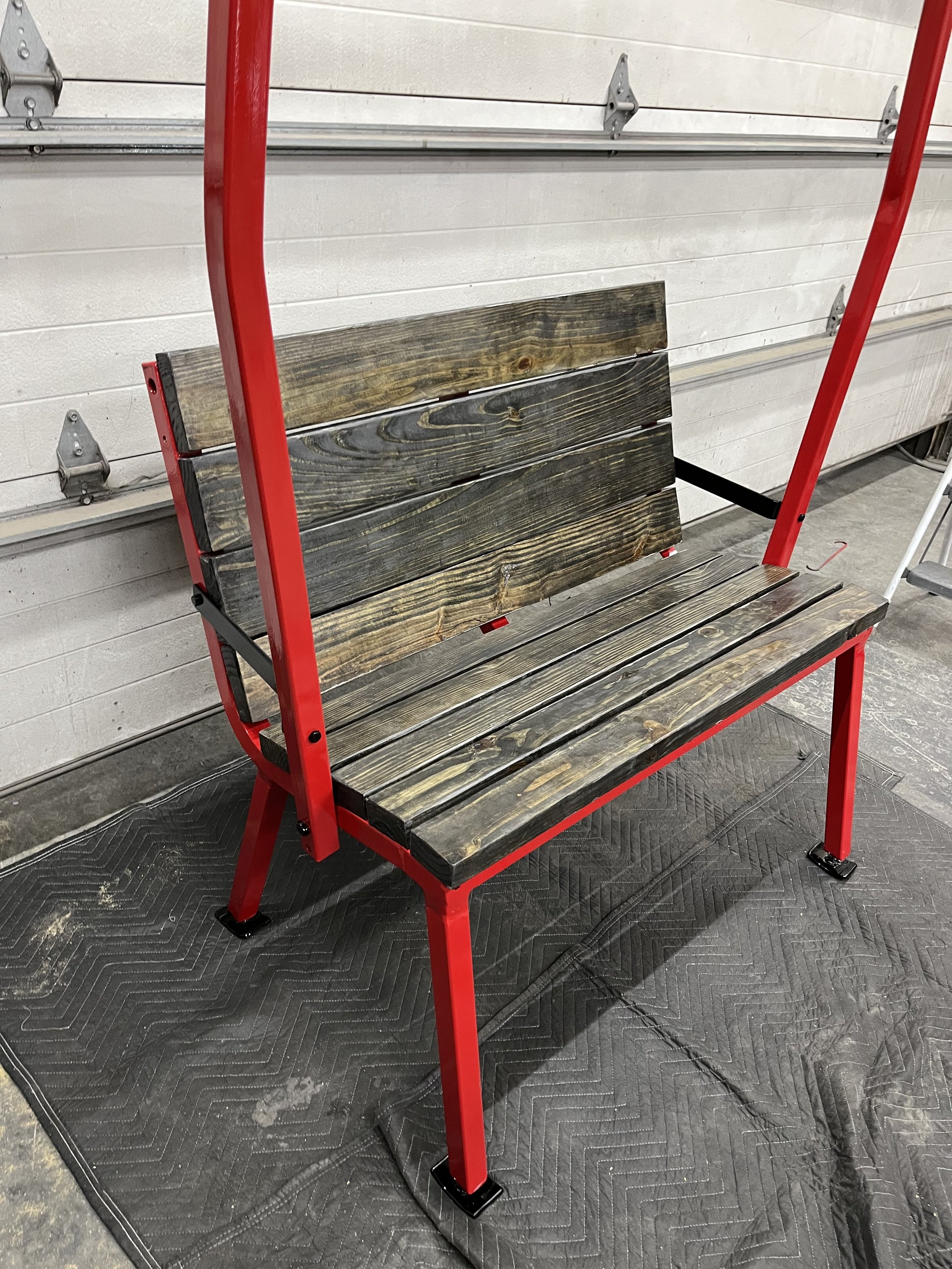 A wooden bench with a red metal frame in a garage, placed on a black padded mat.