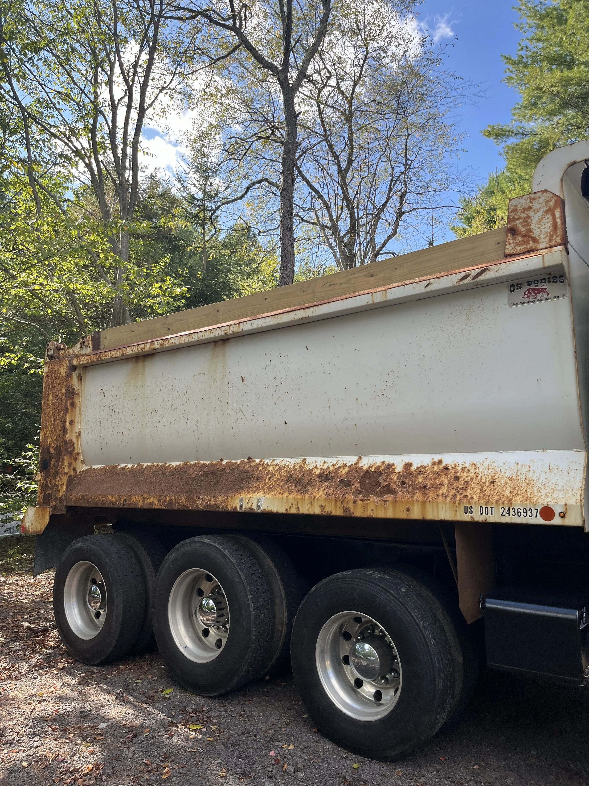 A rusted white dump truck with three wheels, parked on a dirt and gravel surface, with green trees and a blue sky in the background.