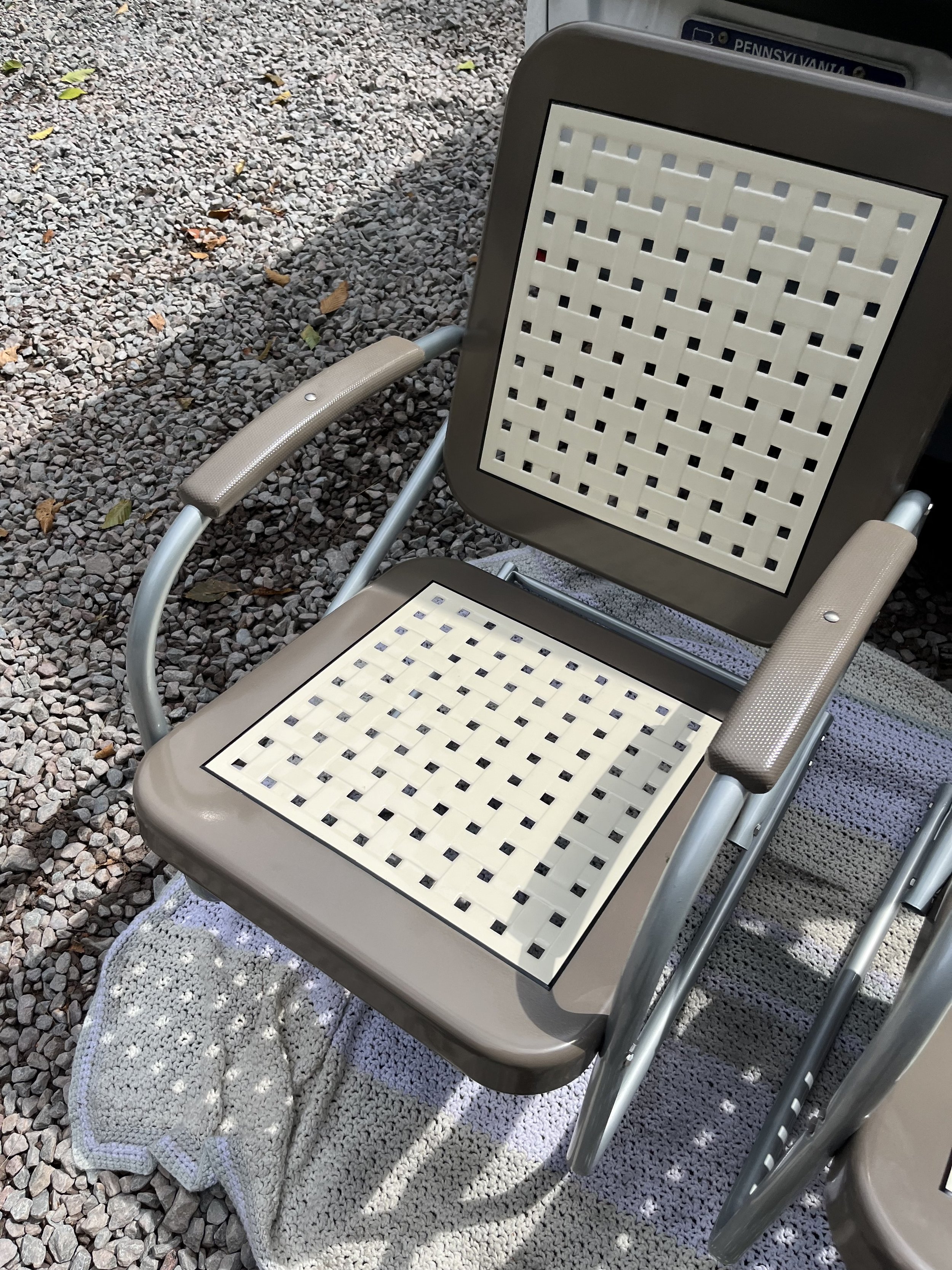 A beige and gray rolling chair with a woven seat and backrest, placed on a white crocheted blanket on gravel ground, with a car partially visible in the background.