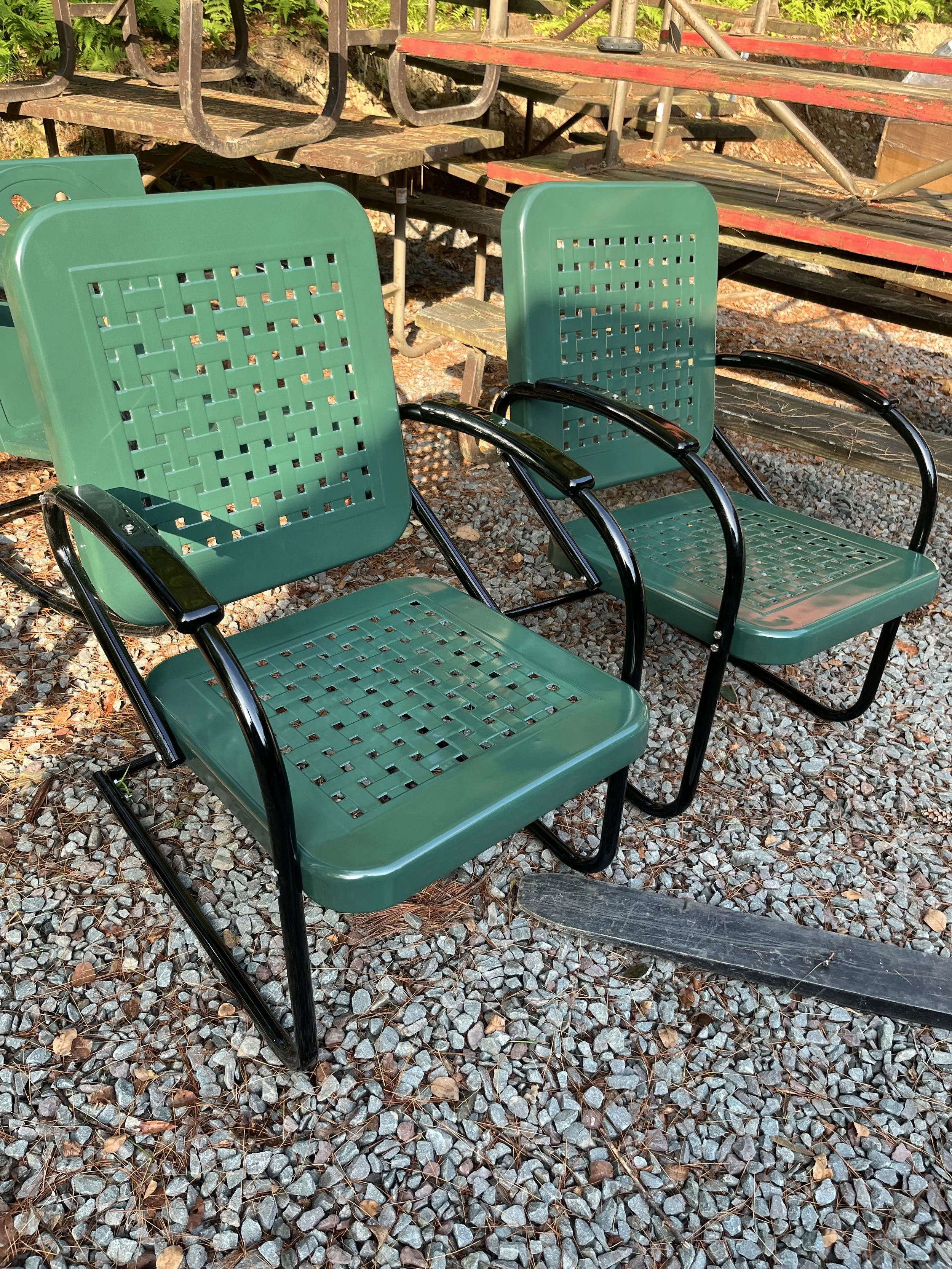 Two green metal outdoor chairs with black armrests placed on gravel ground, with wooden bleachers in the background.