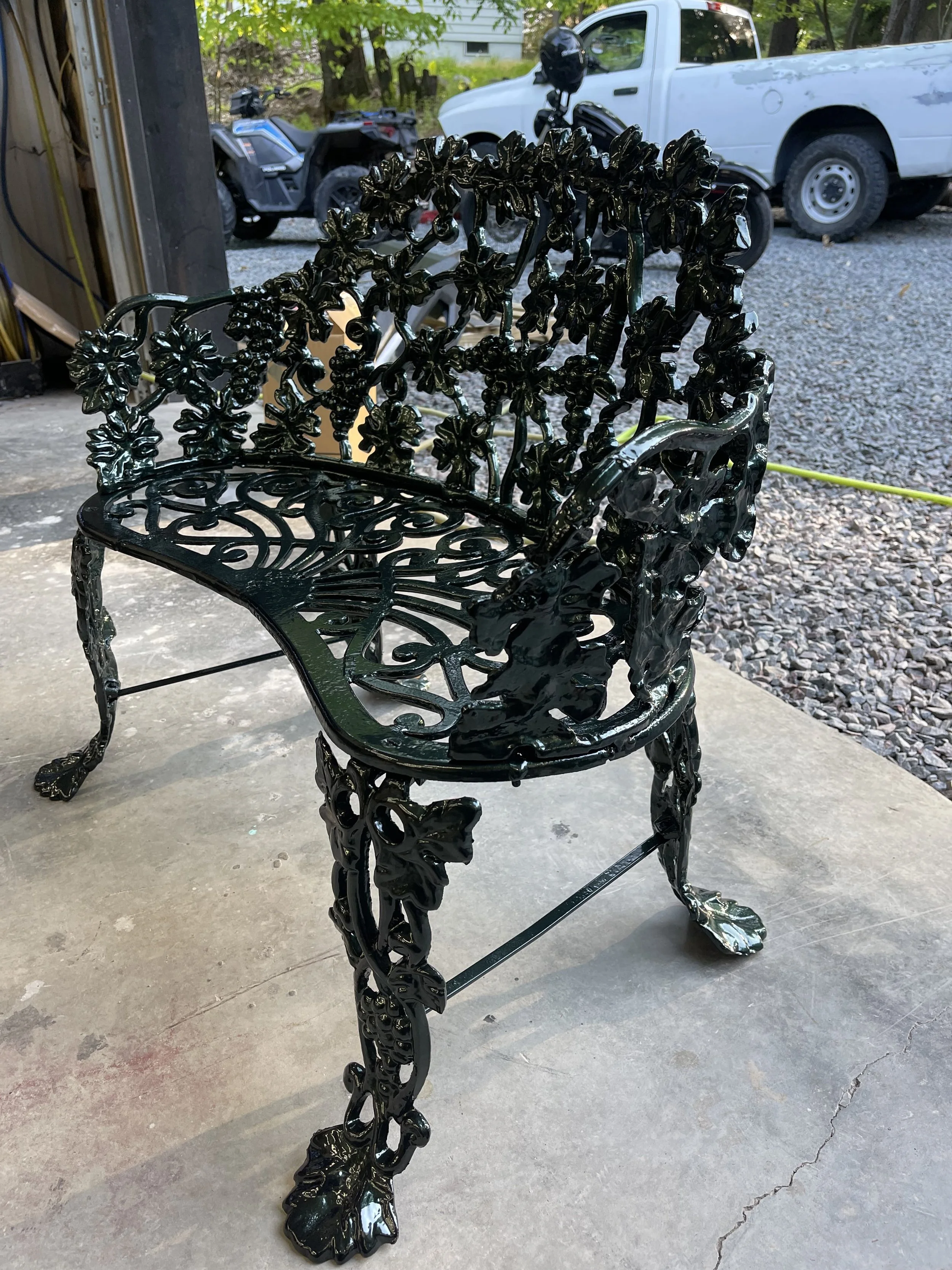Black ornate cast iron garden bench with floral and leaf patterns, set on a concrete floor in a garage or workshop with vehicles and trees in the background.