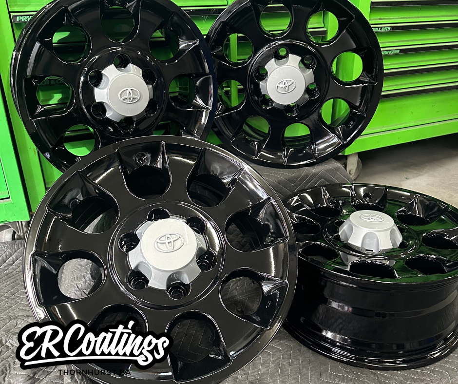 Four black and silver Toyota wheel rims with hubcaps resting on a gray padded work surface with a green tool chest in the background.