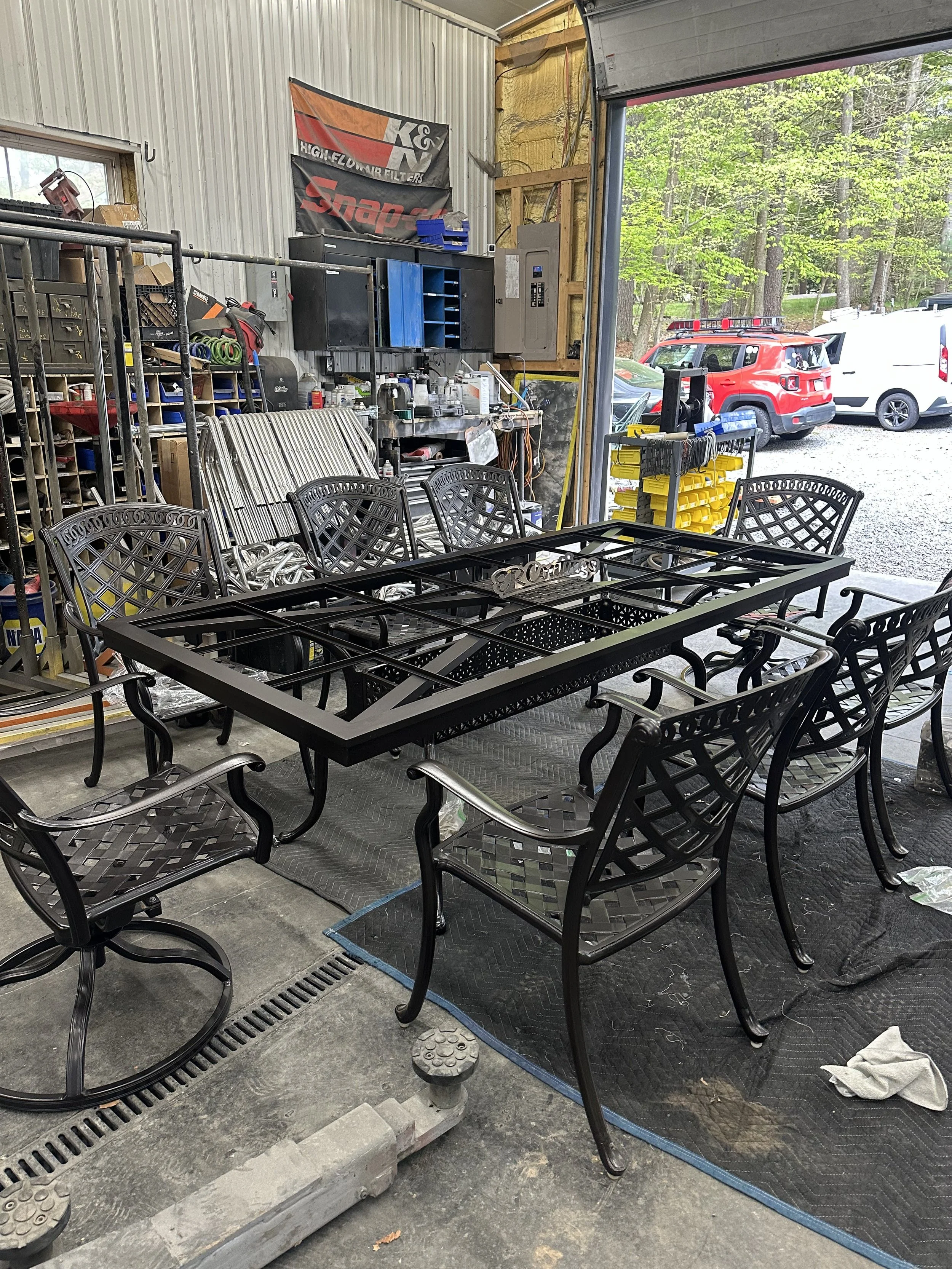 Indoor workshop with patio furniture, including a rectangular glass-top table and metal chairs, with tools, storage shelves, and automotive parts in the background, near an open garage door showing parked vehicles outside.