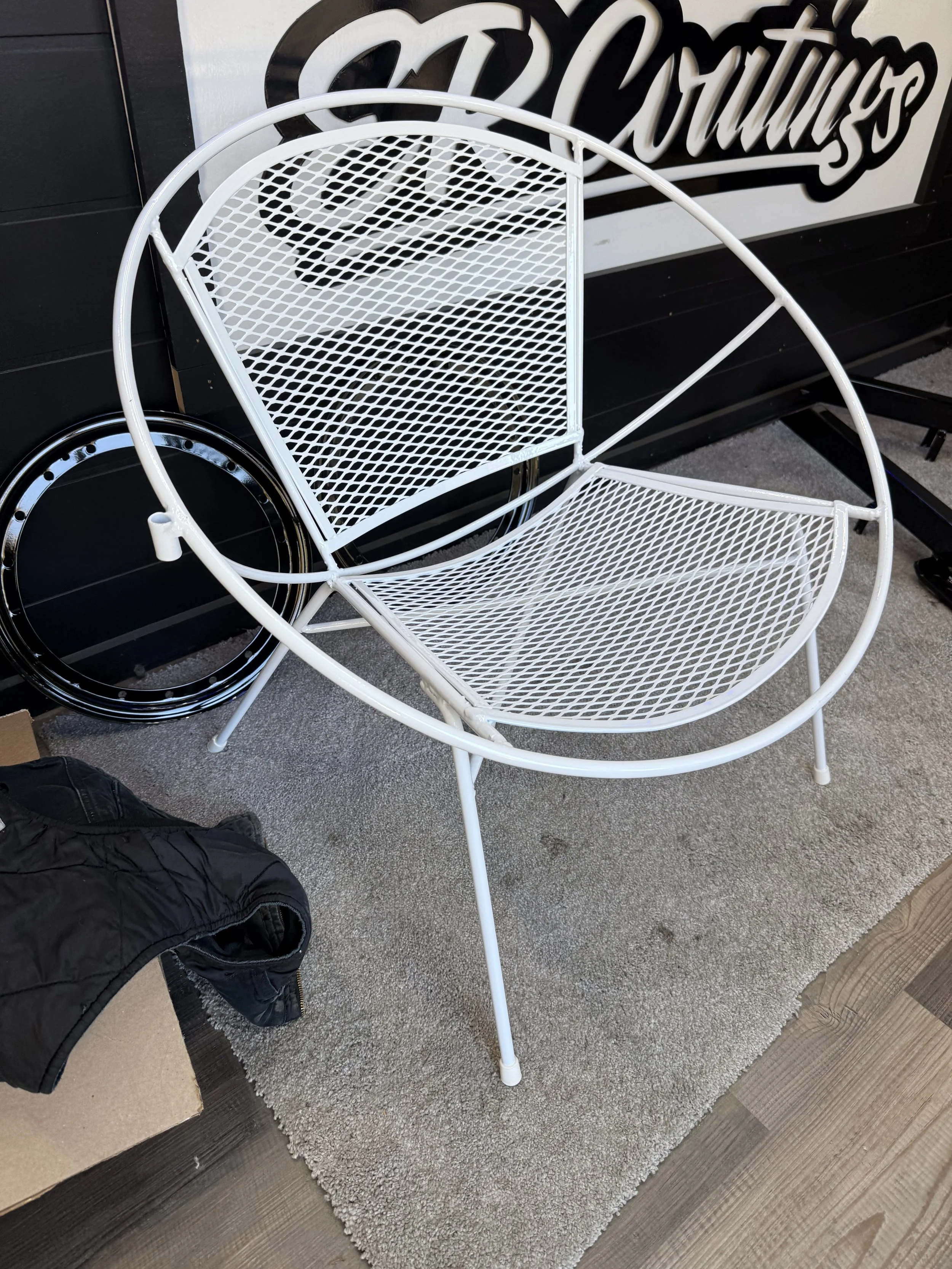 White metal outdoor chair with circular shape and mesh seat and backrest, placed on a beige carpet.