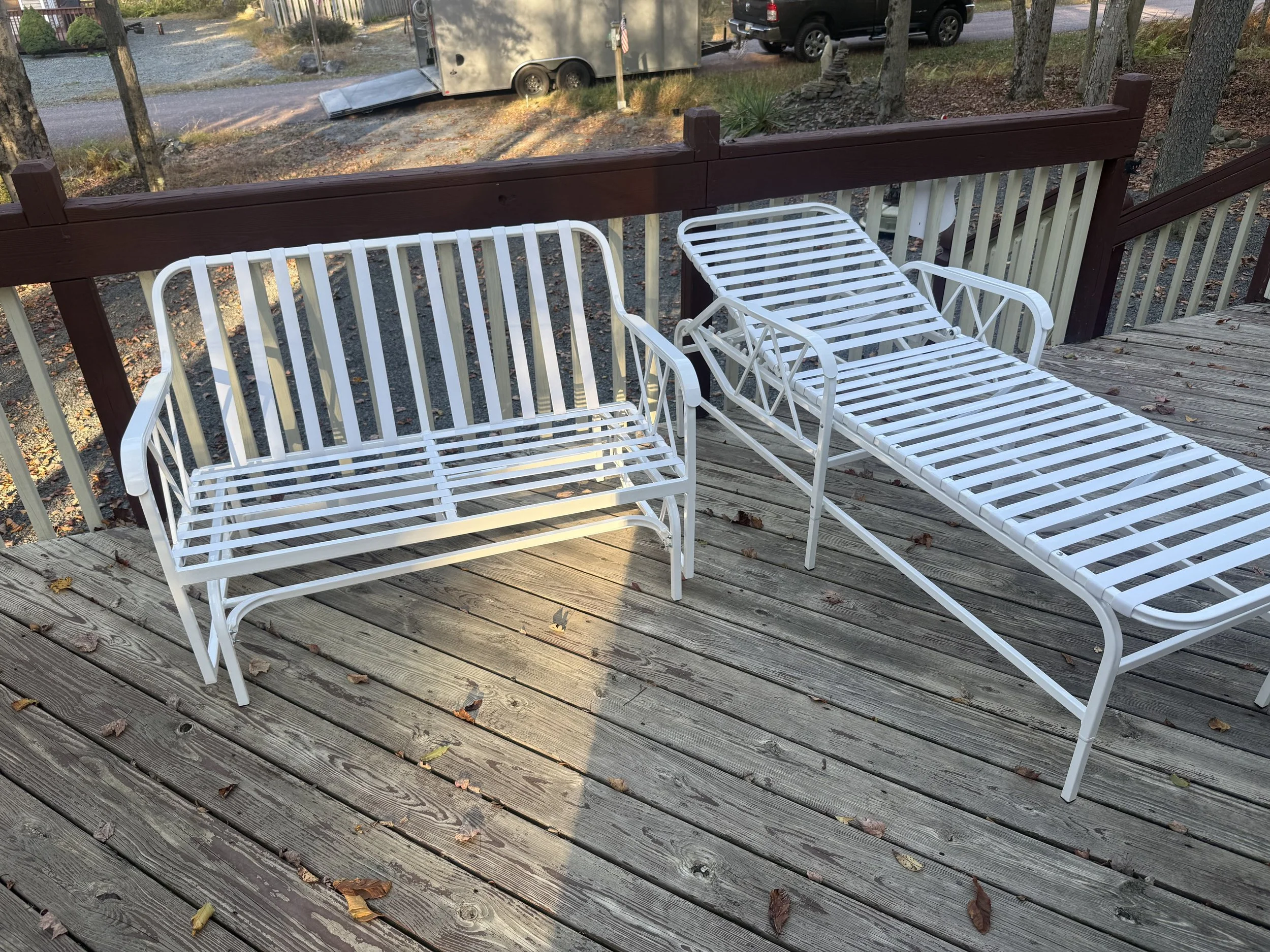 A wooden deck with a white metal bench and a white metal lounge chair, both with slatted seats and backs. The deck has leaves scattered on it, and a red-brown railing surrounds the deck. There are trees in the background and a trailer and vehicles vi