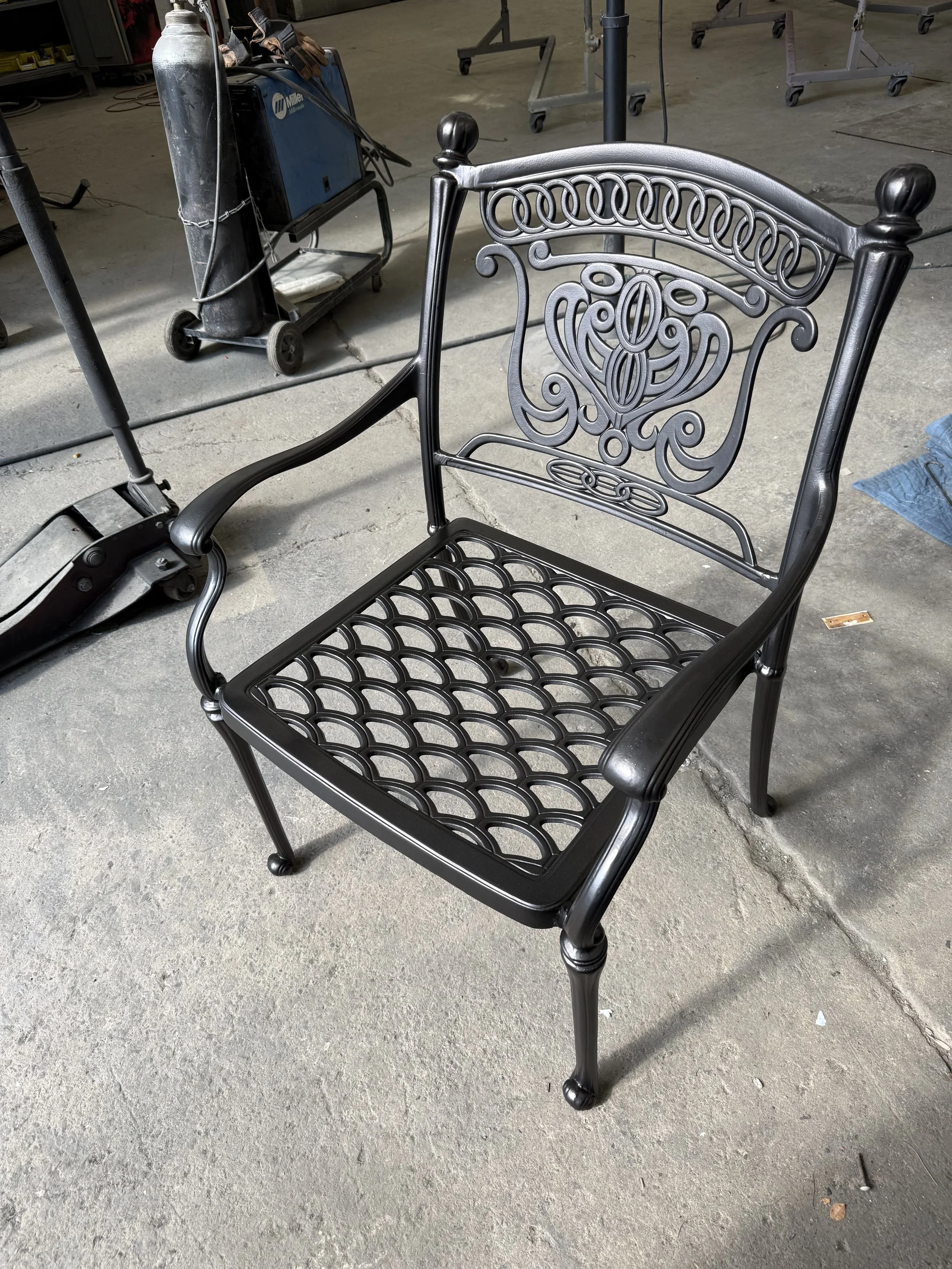 Black metal garden chair with ornate backrest and lattice seat, on a concrete floor in a workshop or garage setting.