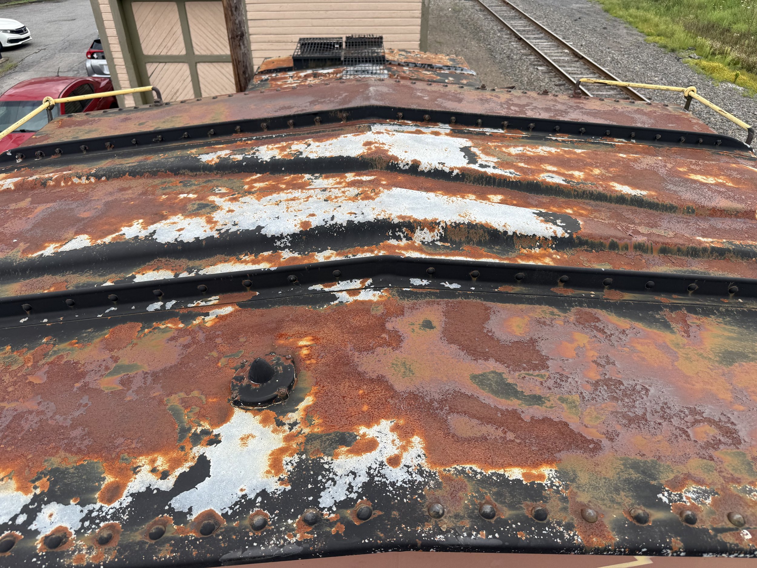 Close-up of a rusty, weathered metal surface with peeling paint and corrosion, possibly on a vehicle or machinery, with screws and a vent visible.