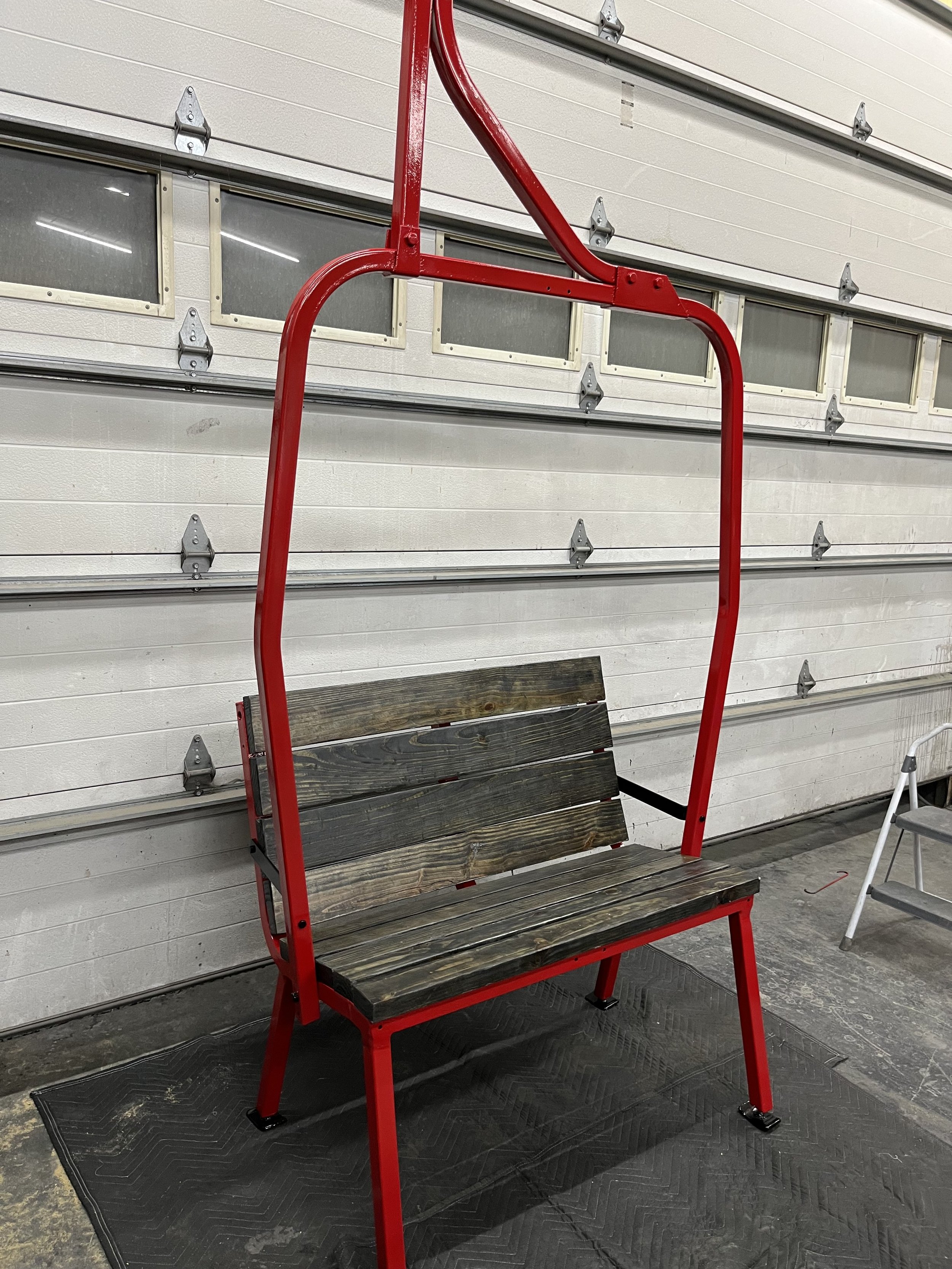 A bench with a wooden seat and backrest, mounted on a red metal frame that extends above the seat to form a support structure.