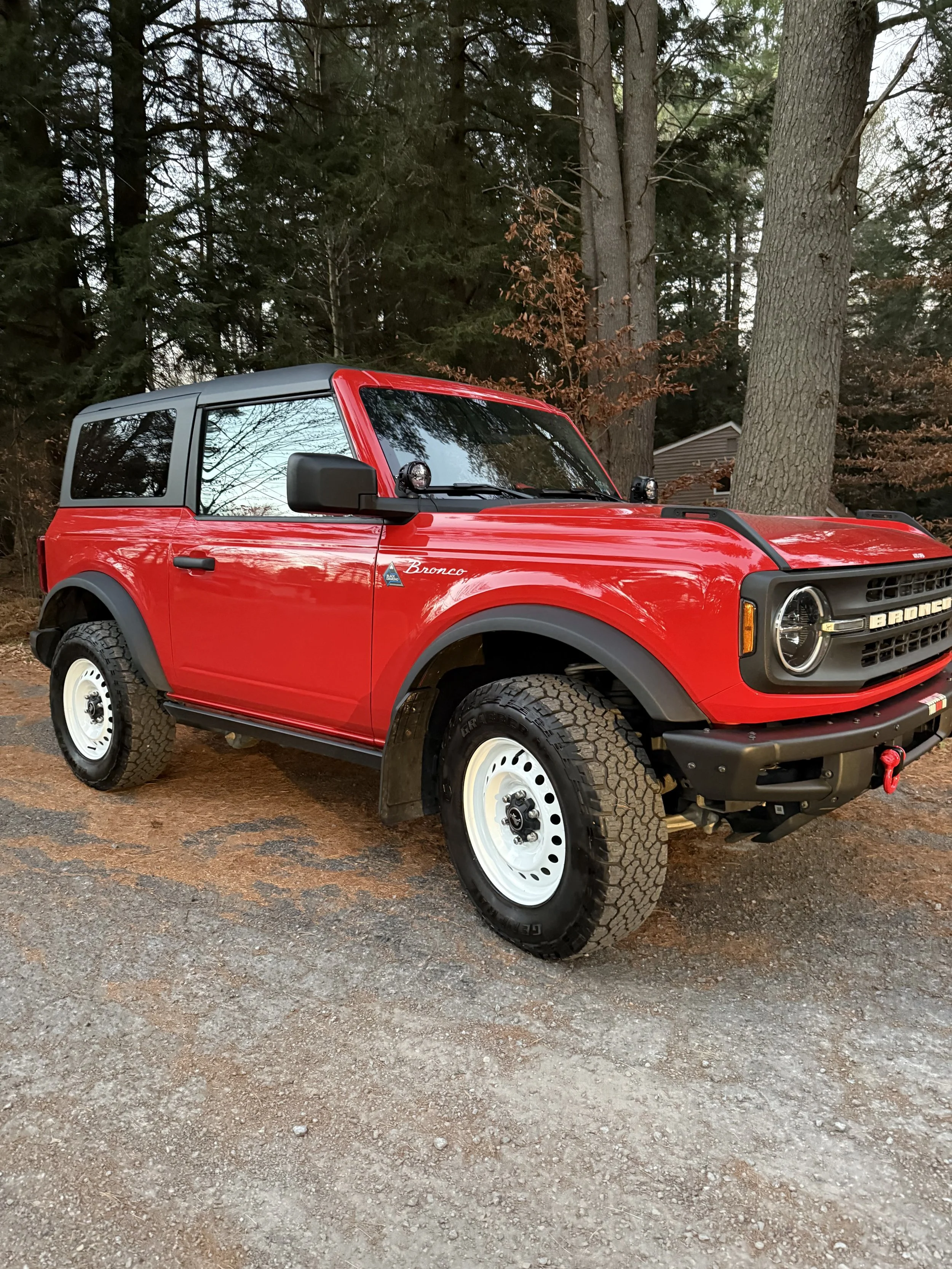 A red Ford Bronco SUV parked outdoors on a dirt surface, with tall trees and a house in the background.
