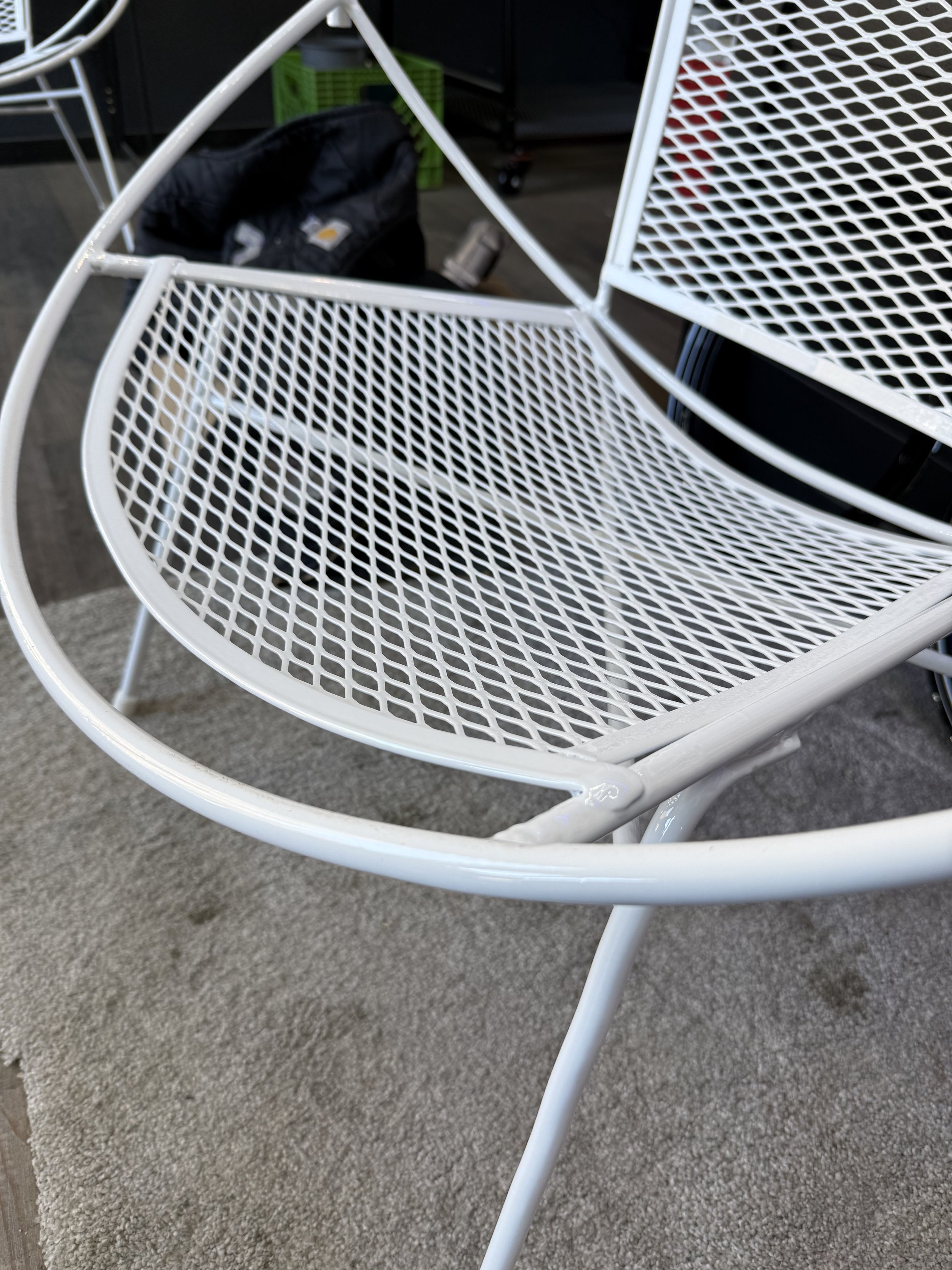 Close-up of a white metal outdoor chair with a mesh seat and backrest, placed on a concrete and carpet surface.