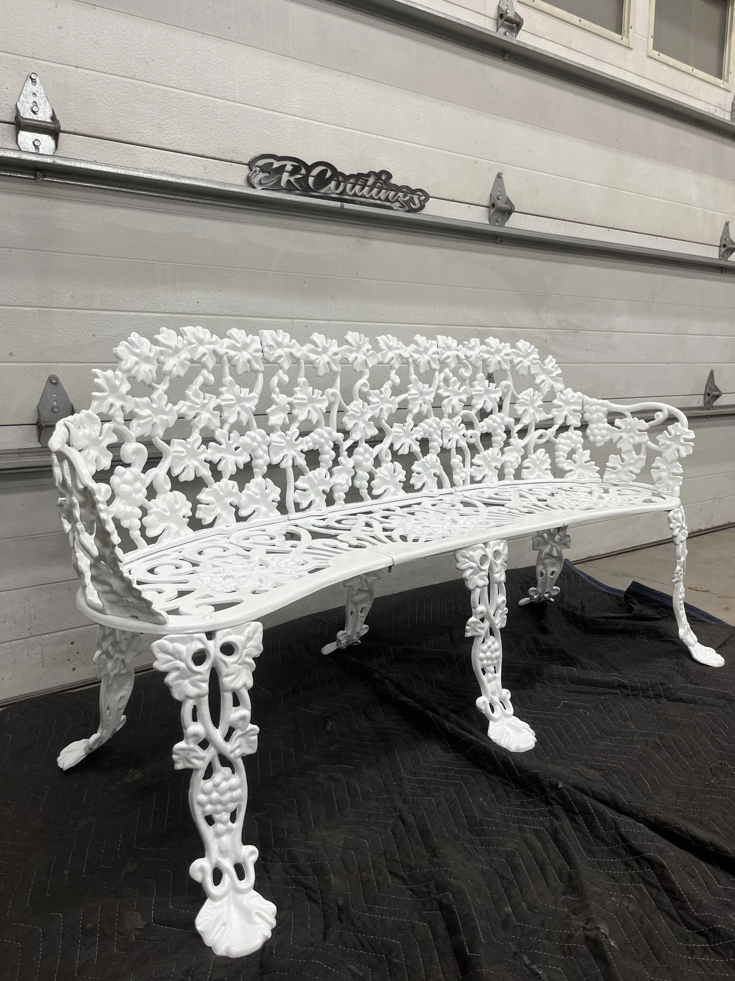 White ornate metal garden bench with floral and vine patterns, placed on a black quilted mat in a garage.
