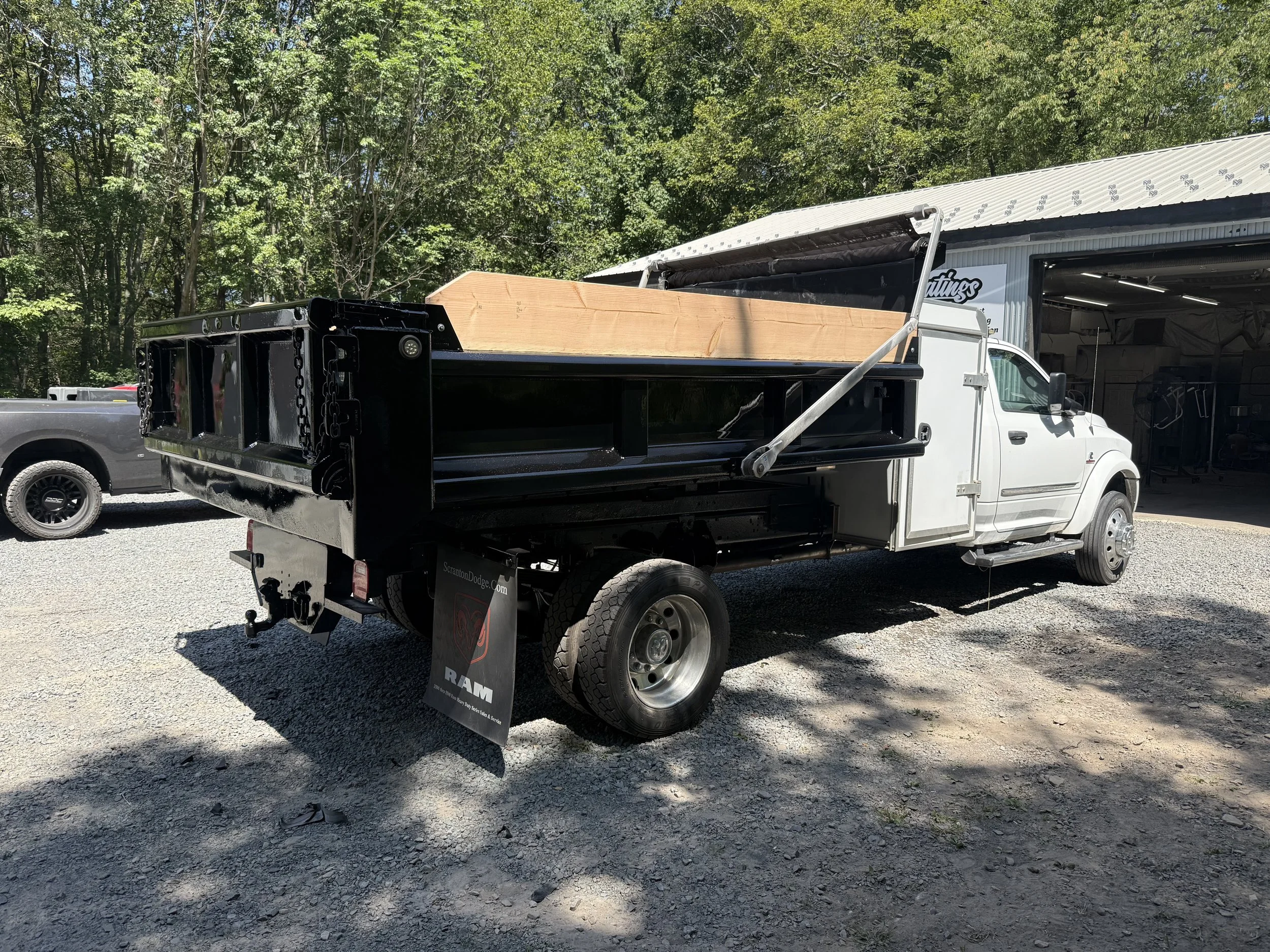 A white flatbed truck with a black bed carrying a large piece of wood, parked outside a garage with a building surrounded by trees.