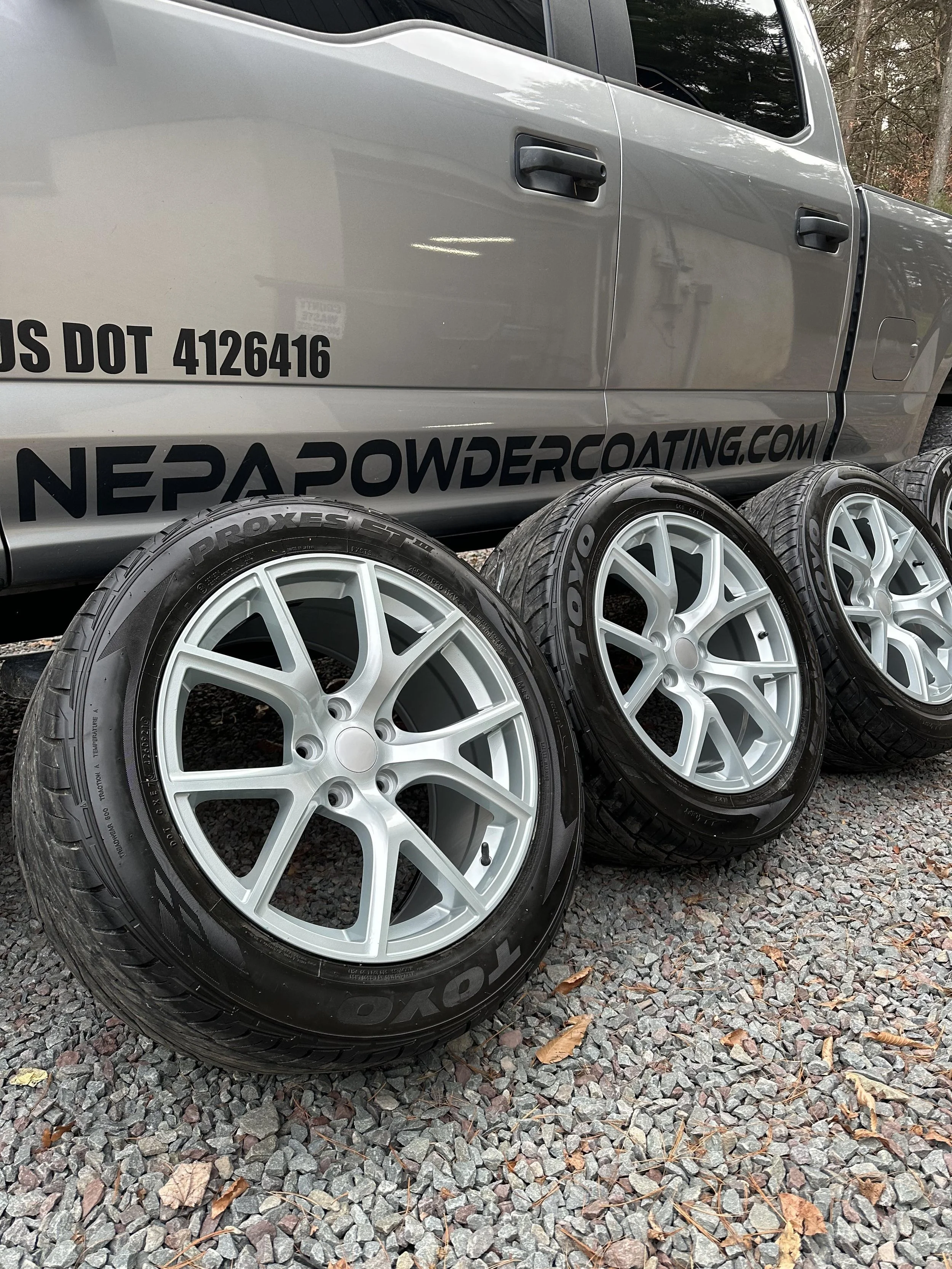 Four silver alloy wheels with Toyo Proxes tires leaning against a gray vehicle with a decal reading 'NEPA POWDER COATING' and a website URL.