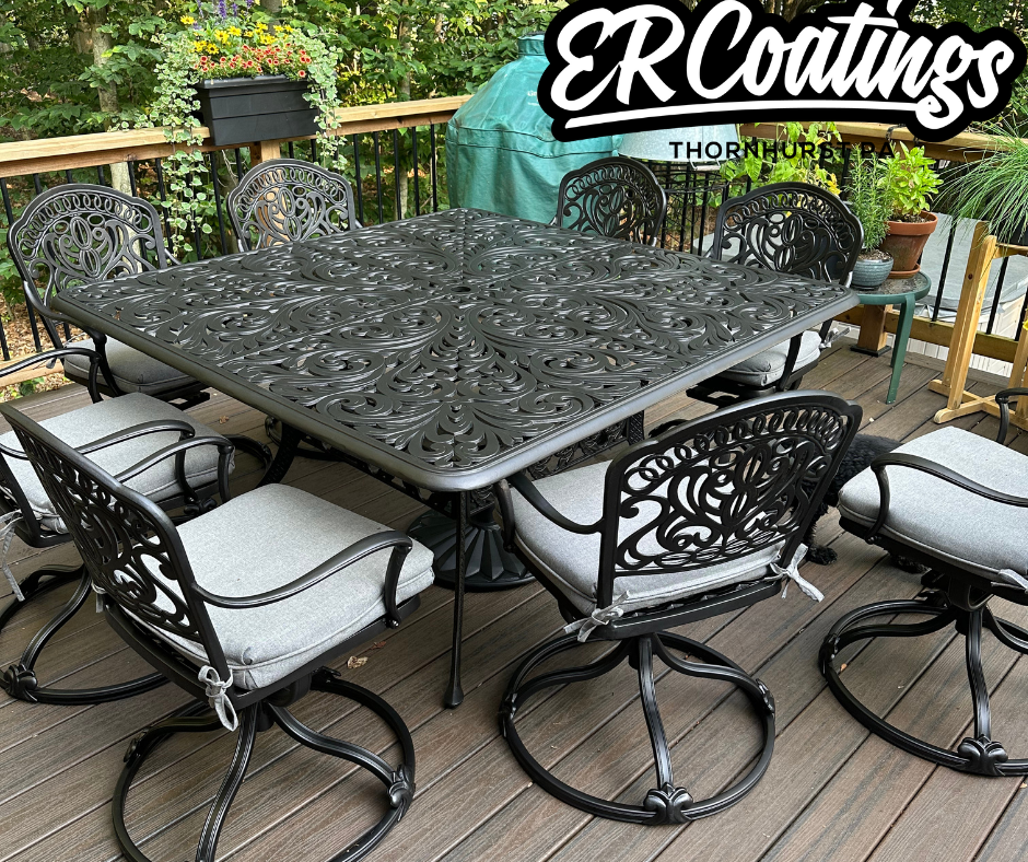 Outdoor patio dining table with six matching chairs featuring ornate black metalwork and light gray cushions. The table has a decorative, intricate metal pattern. The patio is surrounded by a wooden railing, greenery, and potted plants, with a covere