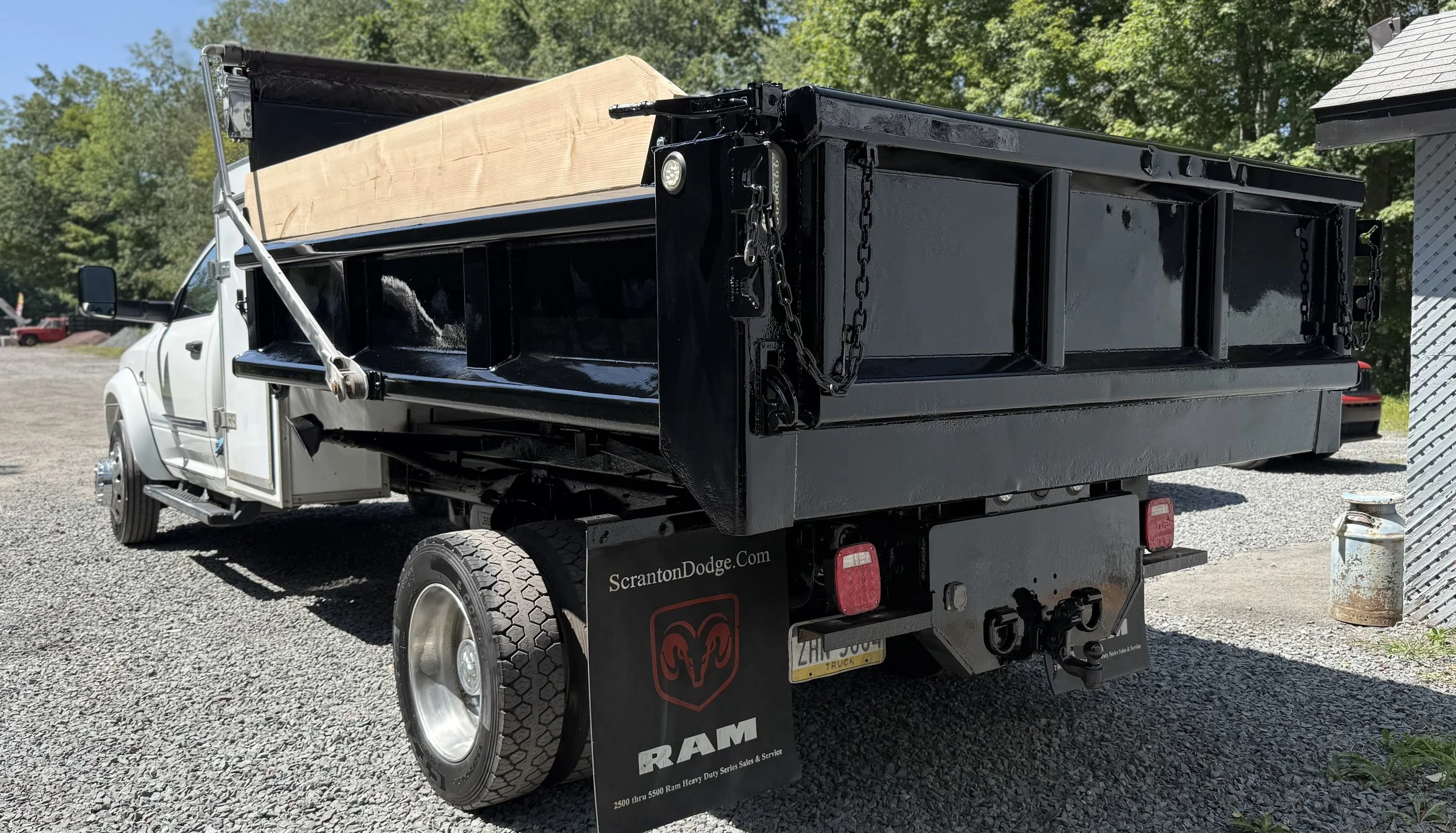 A black flatbed truck with a white cab parked on a gravel lot with trees in the background. The truck has a wooden plank on top of the bed and a DOT tag hanging below. There is a website URL on the mud flap reading "ScrantonDodge.com" and a RAM logo.