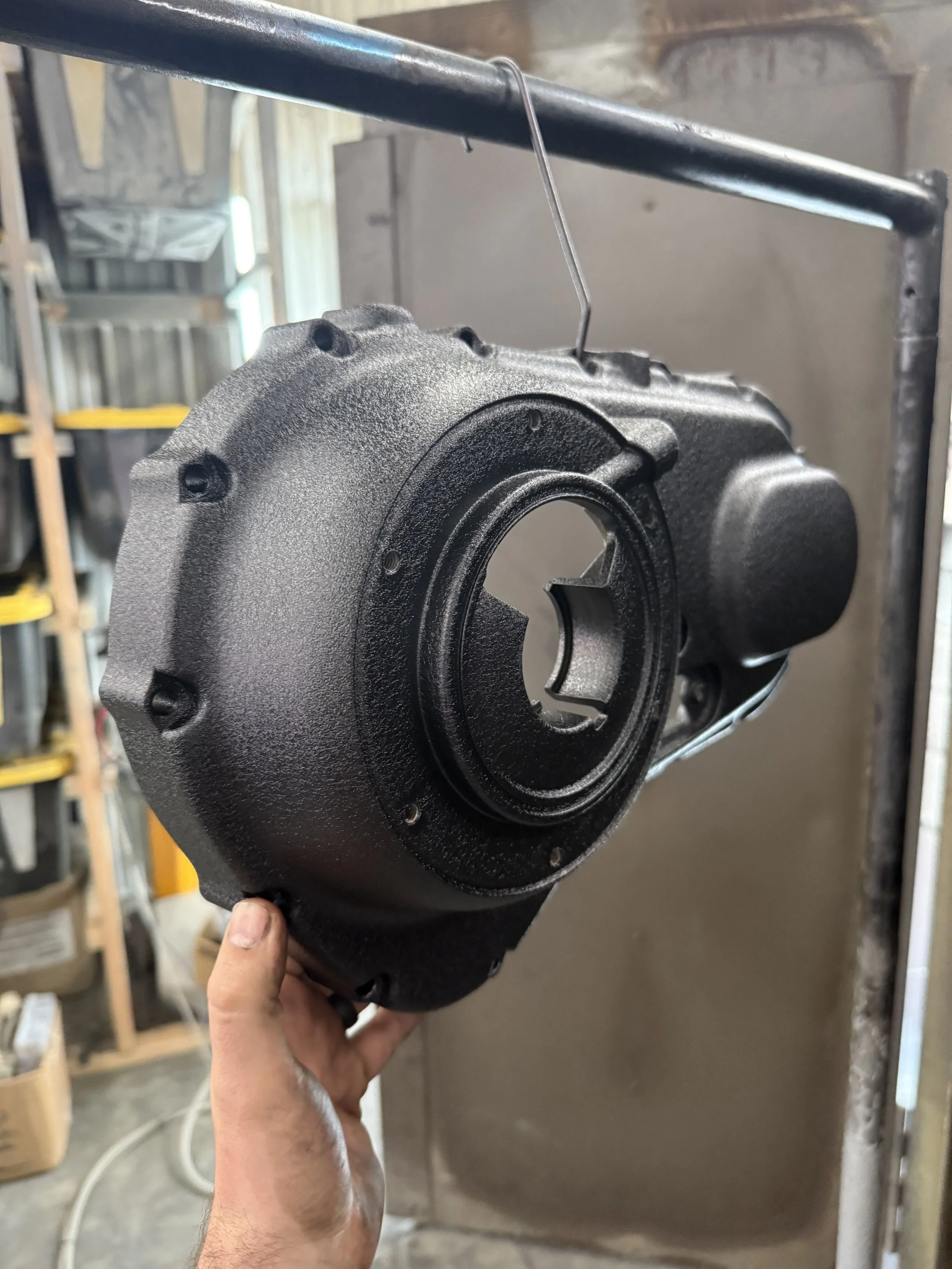 A person holding a black mechanical part, possibly an automotive transmission component, hanging on a metal hook in a workshop setting.