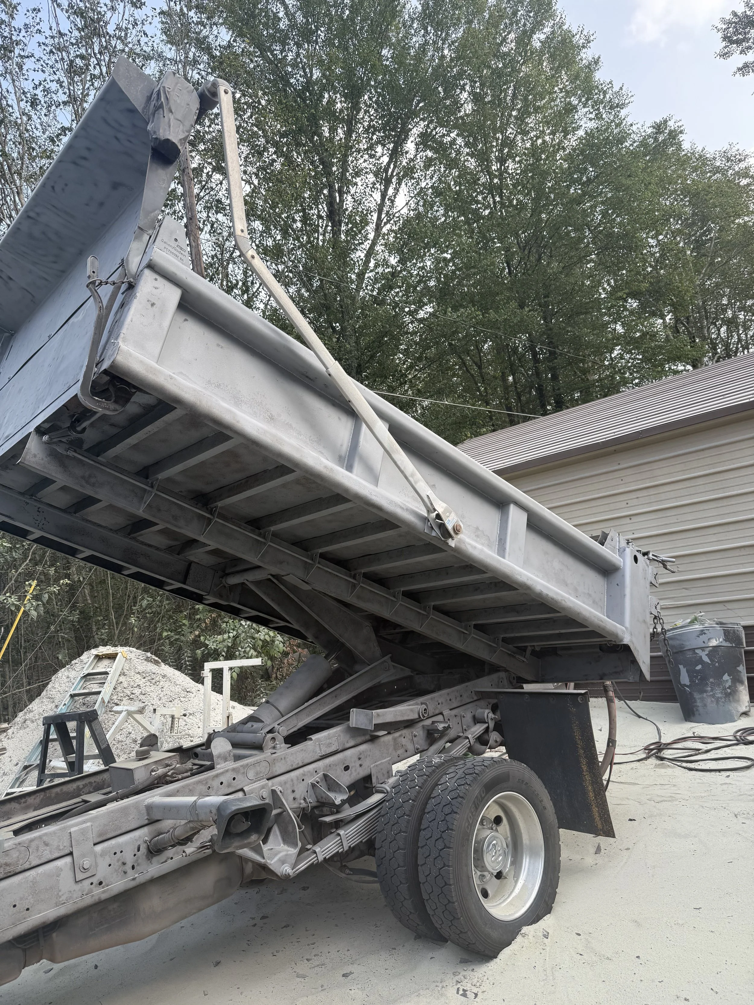 A metal flatbed truck with a partially tipped bed, parked on a concrete surface next to a beige building with a sloped roof, surrounded by trees and construction materials.