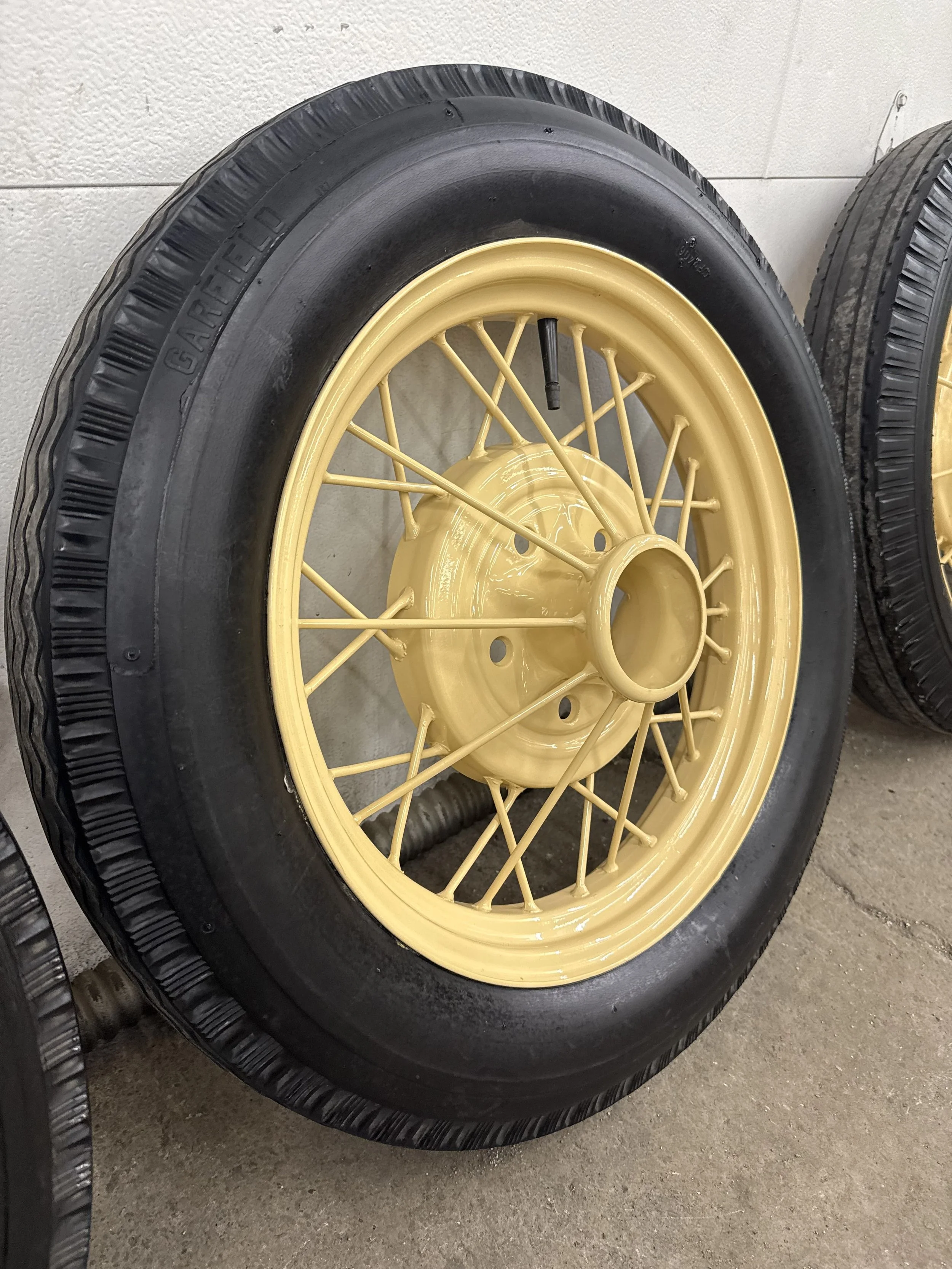 Vintage yellow wire-spoke wheel with a black tire mounted on it, leaning against a light-colored wall.