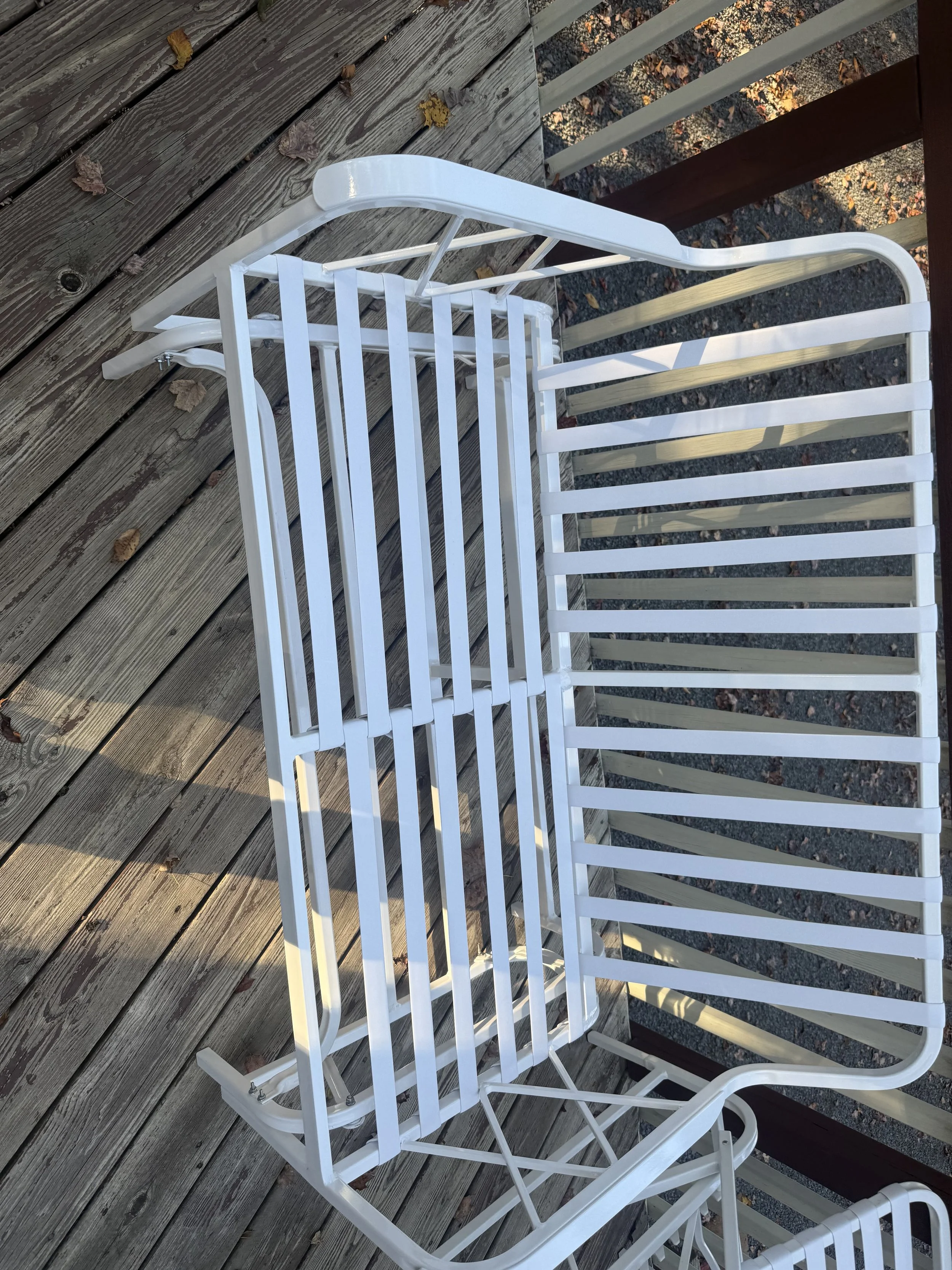 A white metal outdoor bench with a slatted seat and backrest, placed on a wooden deck with fallen leaves.